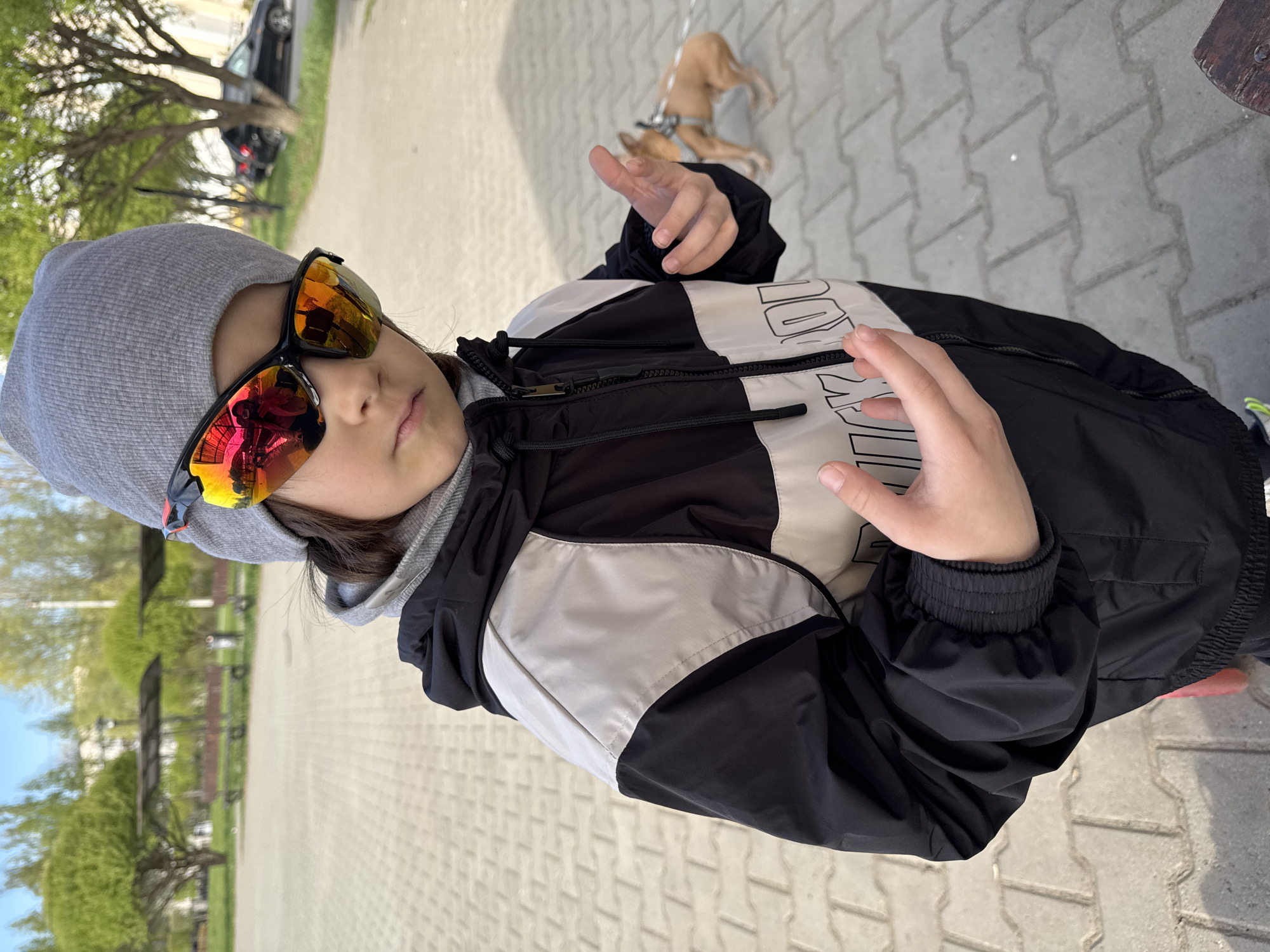 A young person wearing sunglasses and a grey hat stands on a paved surface. They are wearing a black and white jacket and holding their hands up. In the background is a dog on a leash. The location is an outdoor area in the city of Chaikovsky, Russia. There are trees and buildings in the background.