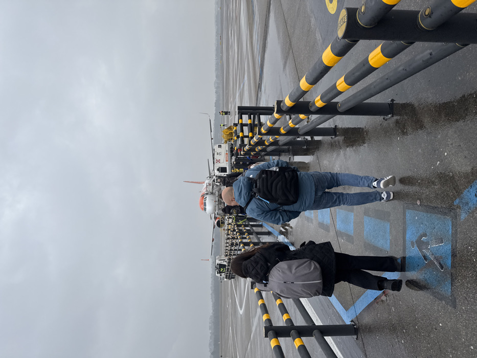 The image depicts an airport tarmac scene on an overcast, wet day at Milan Malpensa Airport in Somma Lombardo, Italy. In the foreground, two individuals walk away from the camera along a designated pedestrian pathway marked with blue symbols of a walking person and arrows. The path is bordered by black vertical posts with two horizontal bars featuring alternating black and yellow stripes.
The person on the left wears a dark, possibly quilted jacket, dark trousers, and a light grey backpack. The person on the right is male, bald, wearing a blue quilted jacket, blue jeans, and dark athletic shoes, carrying a black quilted backpack with light blue straps.
In the mid-ground, an EasyJet aircraft, identifiable by its white fuselage and orange tail with the airline's logo, is parked on the tarmac. A mobile passenger stair is connected to the aircraft's front left door. Various ground support equipment surrounds the aircraft, including a pushback tug near the nose and other service vehicles, one with a yellow hose or cable, towards the right. Several indistinct figures, some appearing to be airport personnel in high-visibility vests, are present around the aircraft. The tarmac is reflective due to moisture, indicating rain. The background shows a distant tree line under a uniformly grey sky.