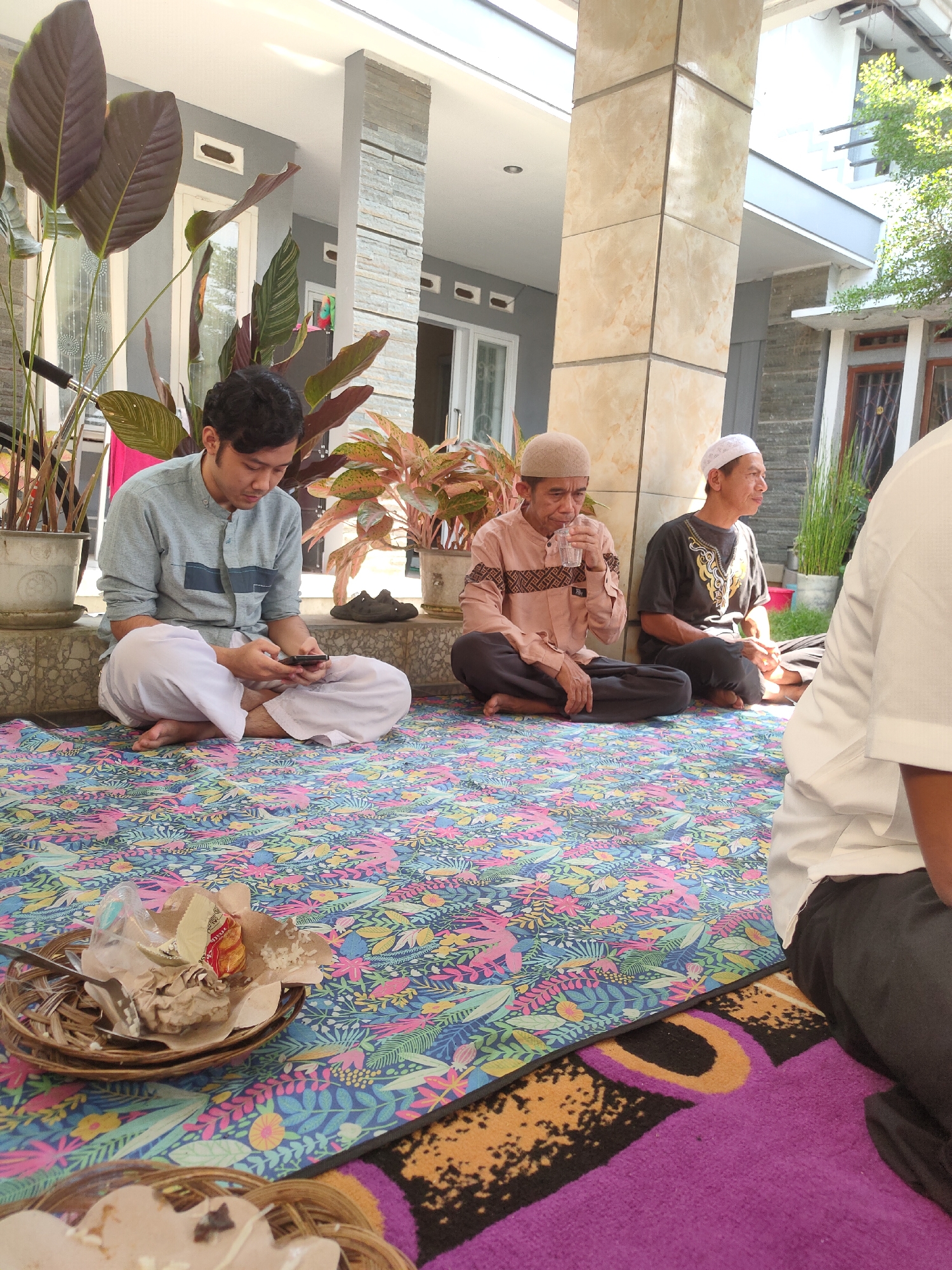 The image depicts a gathering of four men sitting on the floor, likely a residential area in Sumedang, Indonesia, judging by the architectural style and vegetation. Two men are seated on a colorful, floral patterned rug, facing left. One man in a light blue shirt and white pants is looking down at his phone. The other, older man, in a peach-colored shirt and dark pants, is drinking from a glass. To their right, another older man in a dark shirt and white cap is looking to the side. A fourth man, mostly obscured and wearing a white shirt and dark pants, is in the foreground on the right. In front of the seated men are woven baskets containing food remnants. The background features a home with stone and tiled columns, large potted plants, and windows. The scene appears to be outdoors, possibly a courtyard or porch, with natural light suggesting daytime.