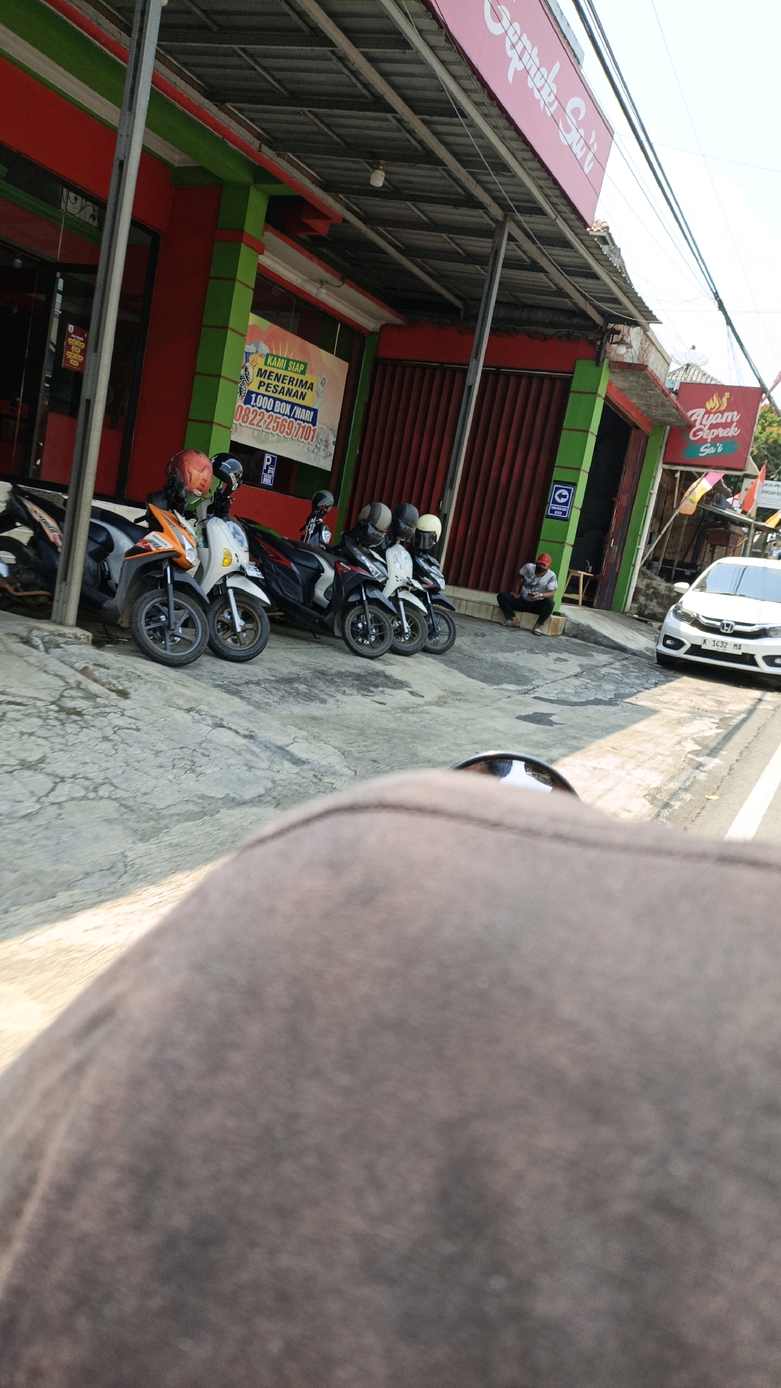 The image depicts a street scene in Kuwasen, Indonesia, featuring a row of parked motorcycles and a white car driving on the road. The motorcycles are lined up along the sidewalk in front of a building with red and green accents. The building appears to be a food establishment, indicated by a sign that reads "Ayam Geprek Suwi". A person is sitting on a small stool near the entrance of the building, and a white car is visible on the right side of the image, driving on the asphalt road. The overall environment suggests a typical urban setting with commercial activity.