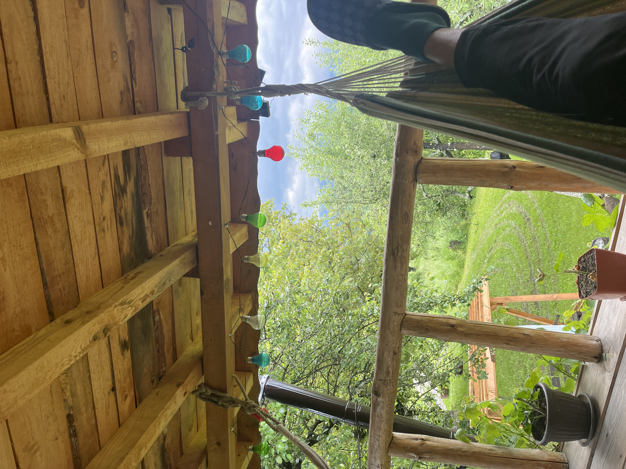 The image shows a partial view from a wooden structure, possibly a porch or veranda, with a hammock hanging from it. A person, likely lying in the hammock, is partially visible in the upper right. A string of colored light bulbs (red, blue, and green) hangs along the wooden beams above. Outside the structure, there is a green, grassy area with trees. Two potted plants are located on the edge of the wooden platform. The image location might be Shopite, Bulgaria.