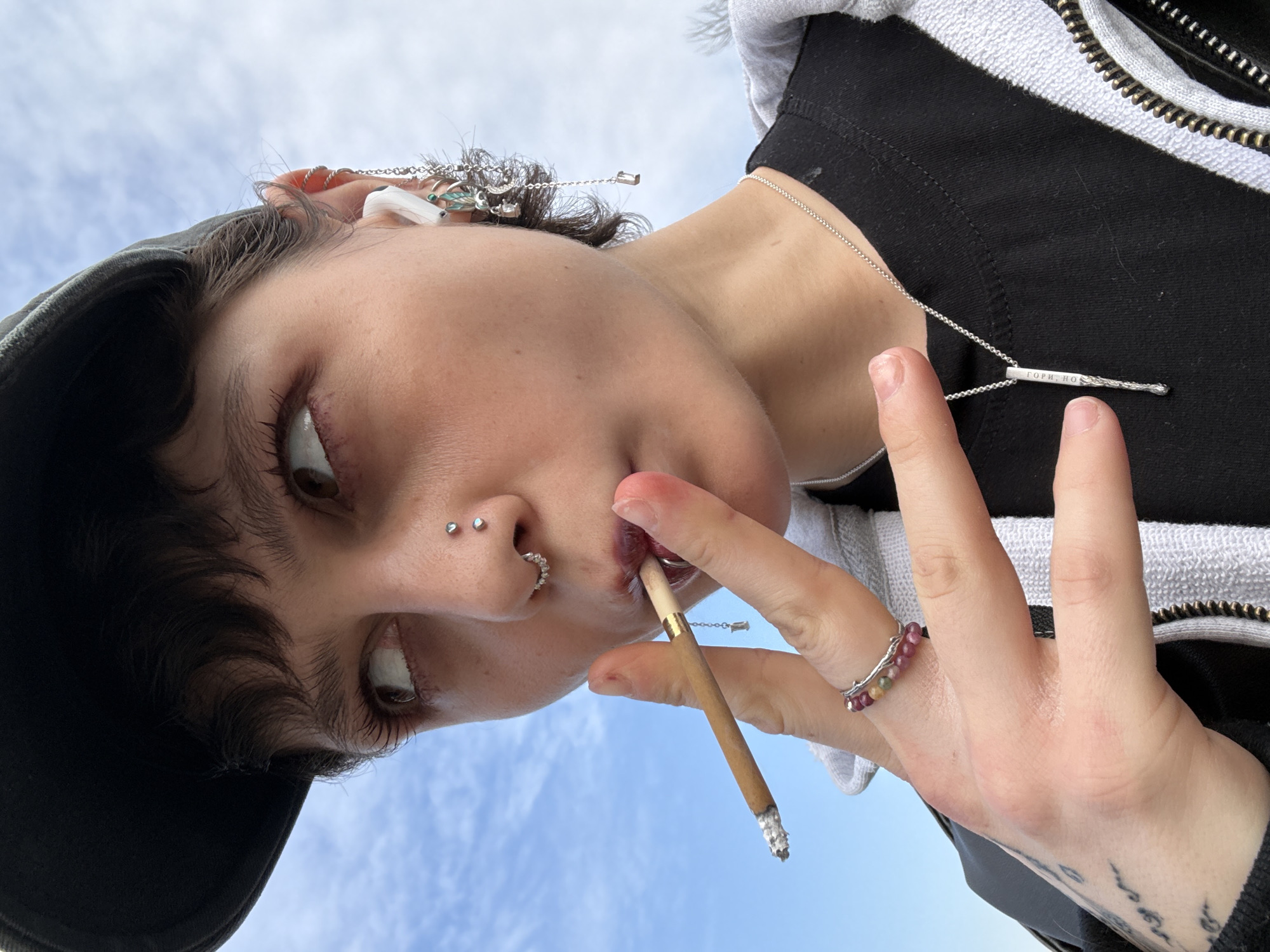 A person with facial piercings (nostril and septum) is smoking a cigarette. The person is wearing a cap and a hooded jacket, along with various pieces of jewelry including a ring, a necklace, and earrings. The background is a light blue sky. The image was taken from a low angle, looking up at the subject. The location is unknown, and there is no information to suggest the scene is in Moscow, Russia.