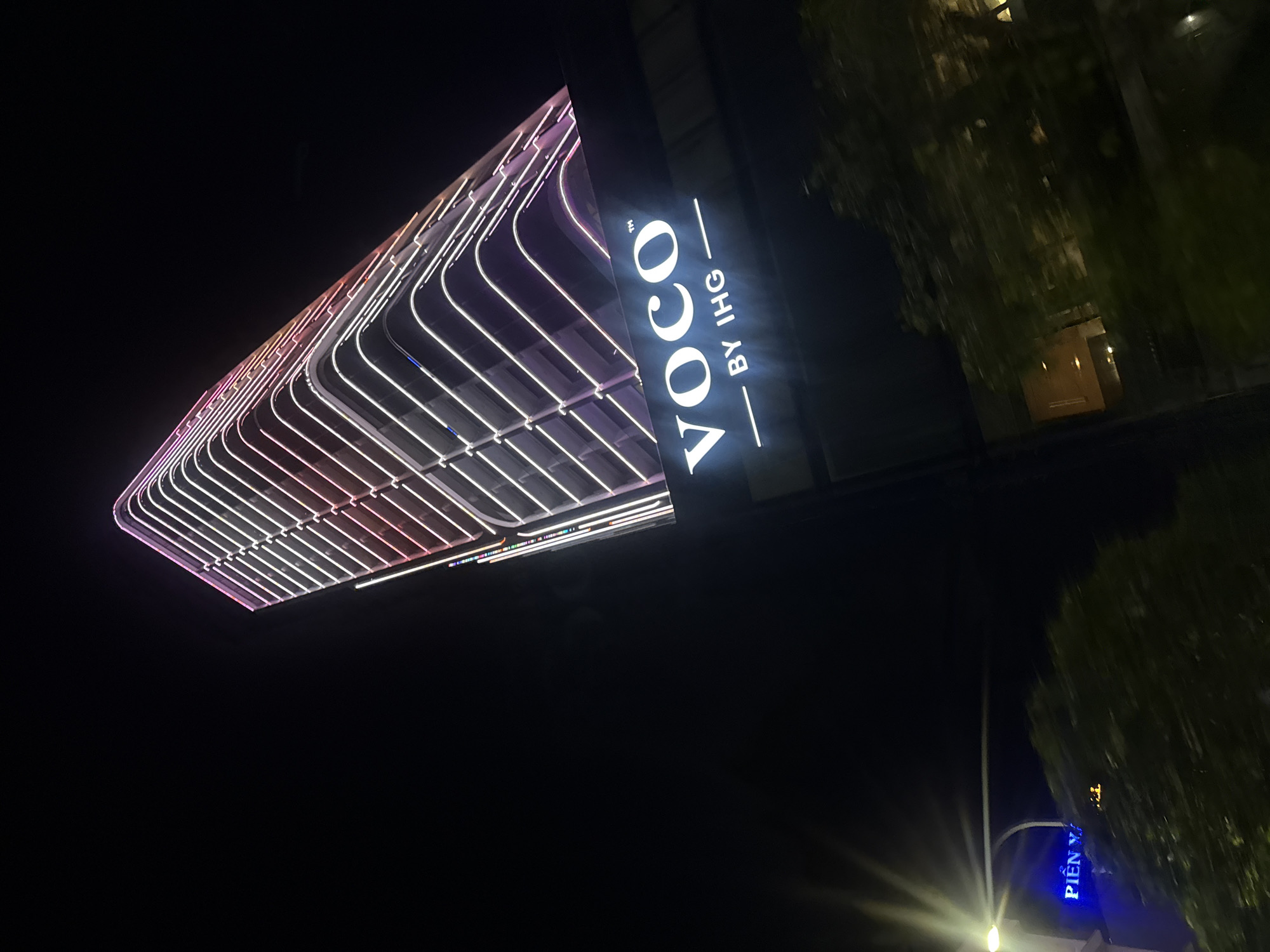 The image shows a modern skyscraper at night in Bac Nha Trang Ward, Vietnam. The building is illuminated with horizontal neon lights that create a striped pattern along its facade. At the base of the building, a prominent sign reads "VOCO BY IHG" in white illuminated letters. Below and to the left of the main building, another sign in blue neon lights says "PIEN Y". Trees are visible in the foreground, partially obscuring the lower portion of the scene and suggesting an urban environment. The sky is dark, indicating it is nighttime.
