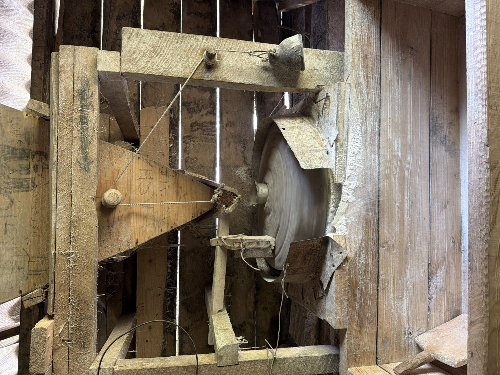 The image depicts a vintage, handmade circular saw enclosed within a wooden frame. The saw blade is spinning, evidenced by the motion blur. The frame appears to be constructed from rough-hewn lumber, showing signs of age and weathering, with dust accumulation and cobwebs. A weight is suspended above the blade, presumably to control the depth of the cut. The surrounding structure is also wooden and appears to be part of a building or shed. The location is potentially in the city of Vakhani, Georgia.