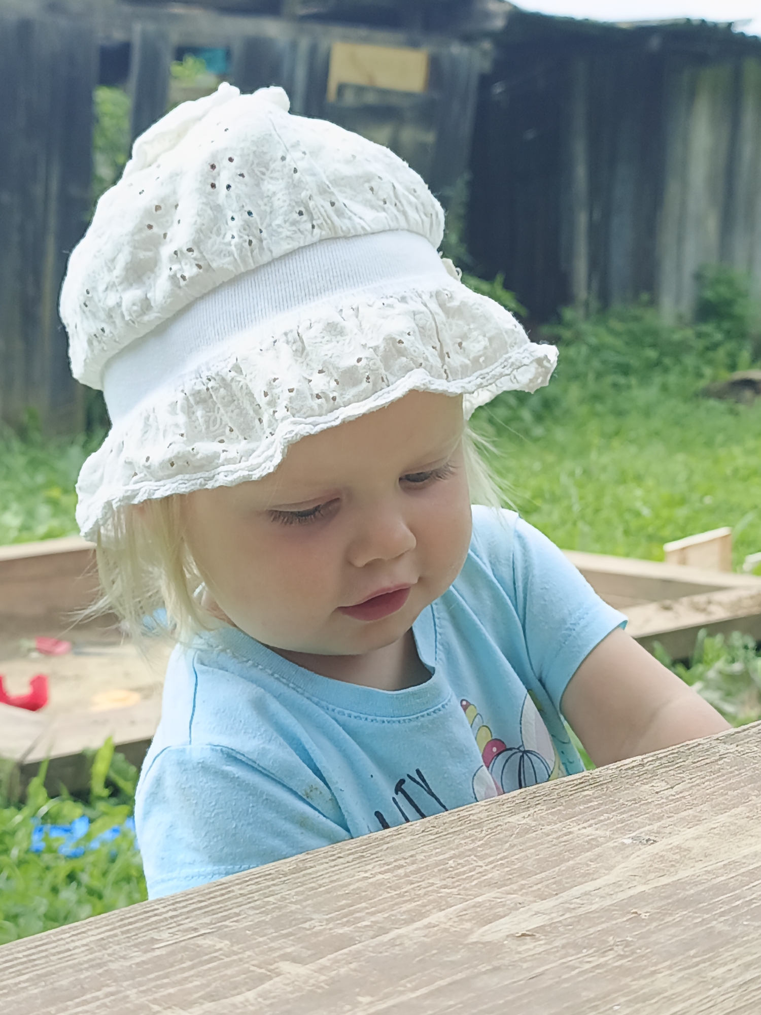 A blonde toddler wearing a white eyelet sun hat and a light blue t-shirt with a unicorn graphic is looking down, likely playing in a sandbox. The sandbox is visible in the background, along with some greenery and a wooden structure. The overall scene suggests an outdoor, possibly backyard, setting in a rural or suburban area. The image has a slightly faded, vintage quality. The location is identified as Medvedyovka, Russia.