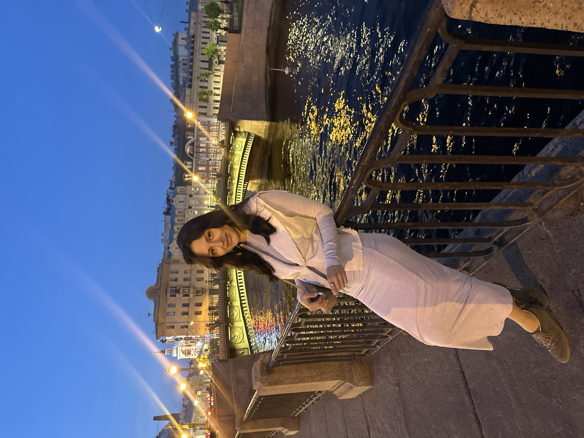 An evening photograph captures a woman posing on a bridge or embankment in Saint Petersburg, Russia, alongside a body of water. The time of day is dusk or early night, characterized by a deep blue sky and the illumination of urban lights.
The primary subject is a woman with dark hair, who is smiling gently and looking towards the camera. She is dressed in light-colored attire, including what appears to be a light grey or off-white jacket or cardigan over a lighter top, a long skirt or wide-leg trousers of similar light hue, and dark sneakers. She is leaning with her left arm on a dark metal railing that separates her from the water below, and she holds a small, light-colored bag or clutch with both hands.
The background features classic European architecture lining the opposite bank of the canal or river. These buildings are lit up, creating bright reflections on the rippling dark water. A decorative bridge structure is visible behind the woman, spanning the waterway. Streetlights emit warm glows, some creating starburst effects in the image. The overall atmosphere is calm and illuminated by artificial light. There is no visible text in the image.