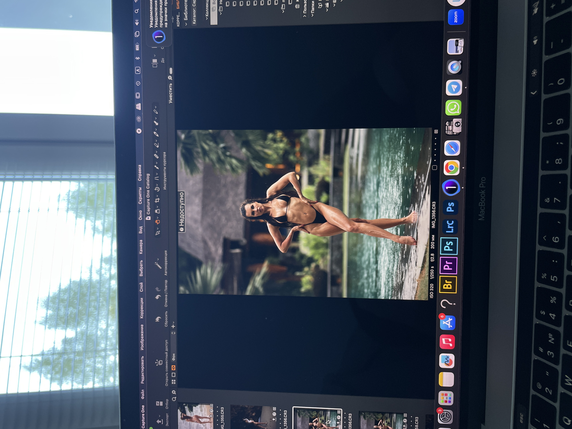 The image is a photograph of a MacBook Pro screen showing the Capture One photo editing software, displaying an image of a woman in a black bikini posing near a body of water. The software interface elements, including the menus (File, Edit, Image, Correction) and toolbar icons (Export, Selection) are visible. Dock icons for various applications, including Safari, Google Chrome, FaceTime, WhatsApp, Telegram, Photos, Adobe Photoshop, Adobe Premiere Pro, and Capture One are seen next to the Finder icon. The MacBook keyboard is partially visible. The location of Kazan, Russia, is not evident in the image. The environment outside the window shows trees and light.