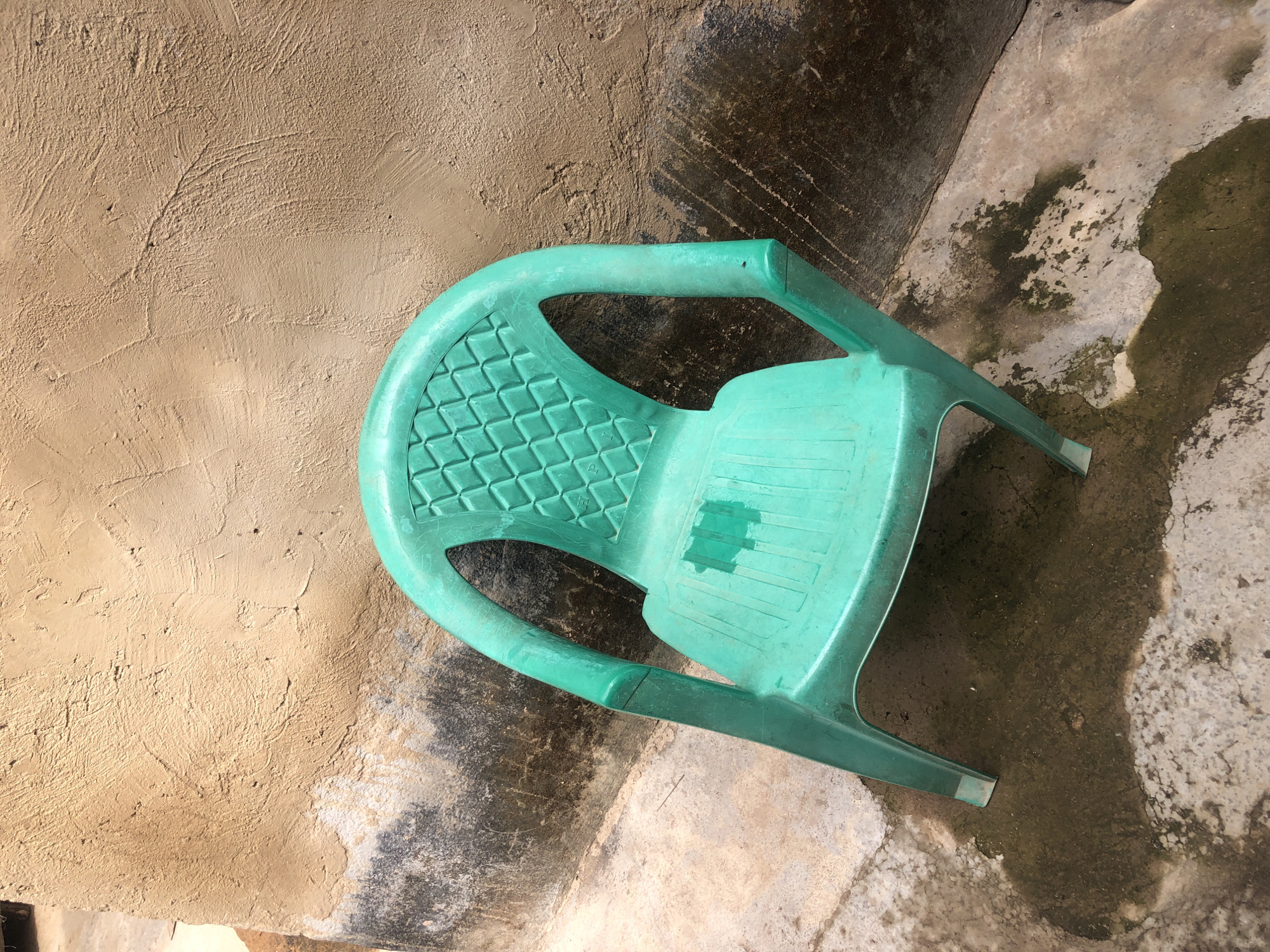 The scene depicts a solitary green plastic chair positioned outdoors in Unknown, Unknown. The chair, a lightweight utility model, is a medium shade of teal or aqua green. Its backrest features a diamond grid pattern, while the seat has parallel raised ridges, with a distinct dark, wet patch visible on its center. One armrest shows a prominent vertical seam line and minor surface scuffs.
The chair is leaning against a textured, light brown or tan wall. The wall surface appears to be rough plaster or stucco, exhibiting an uneven, trowel-marked finish. Approximately one-quarter of the wall's lower section is discolored by a dark, irregular band of moisture and grime, transitioning sharply from the drier upper portion.
The ground beneath the chair is a concrete or paved surface, irregularly wet and stained with dark patches, suggesting pooled water, possibly mixed with dirt or algae. These wet areas are most concentrated around the chair's feet and spread outwards. Visible cracks and varying textures are present in the concrete. The lighting is bright and direct, casting subtle shadows and highlighting the textures of the wall and ground. No individuals, animals, or other significant objects are present in the immediate vicinity.