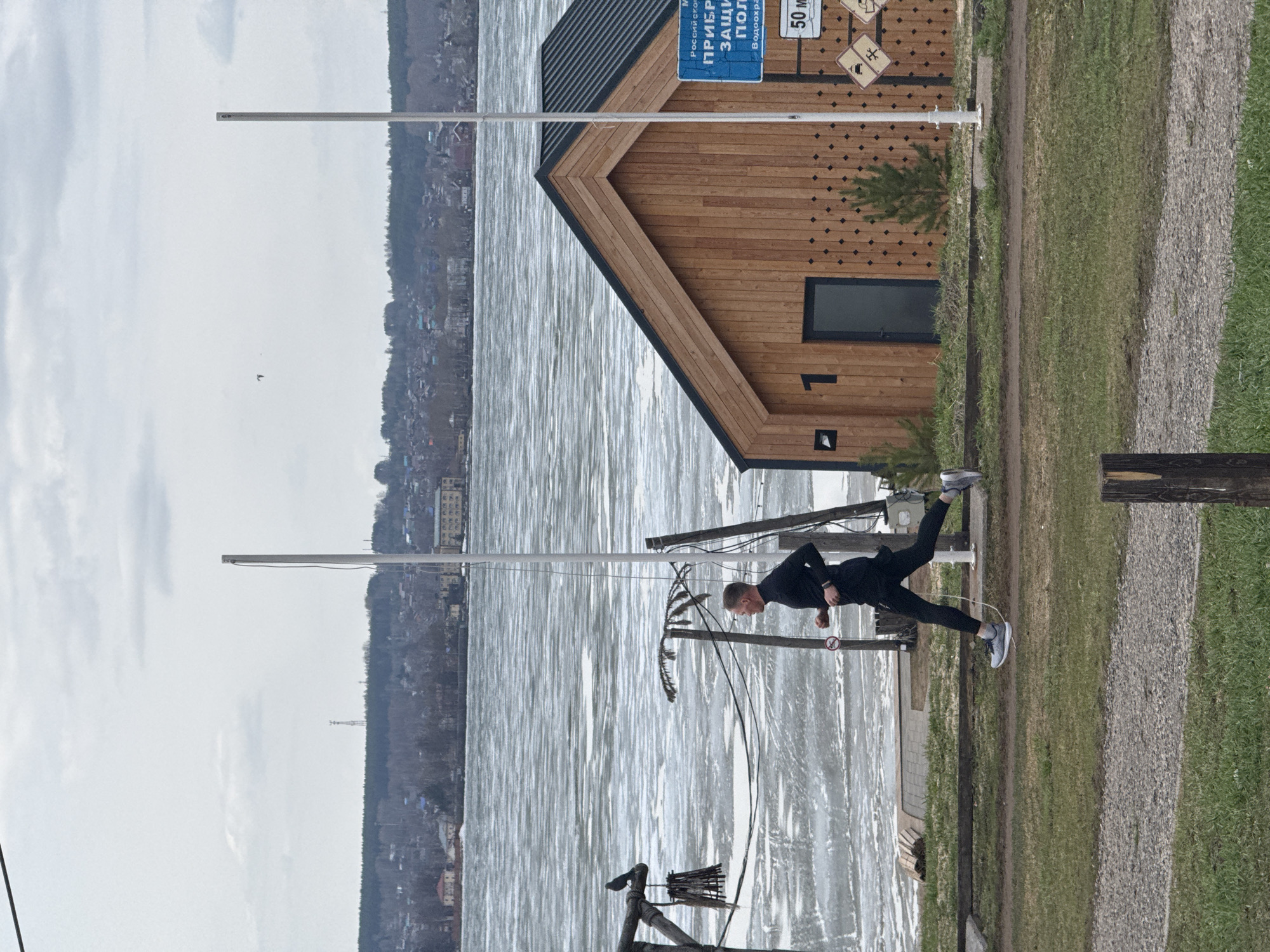A man in dark athletic clothes is balancing on one leg on a platform next to a body of water in Votkinsk, Russia. A wooden building is adjacent to him. A town is visible across the water. The sky is overcast.