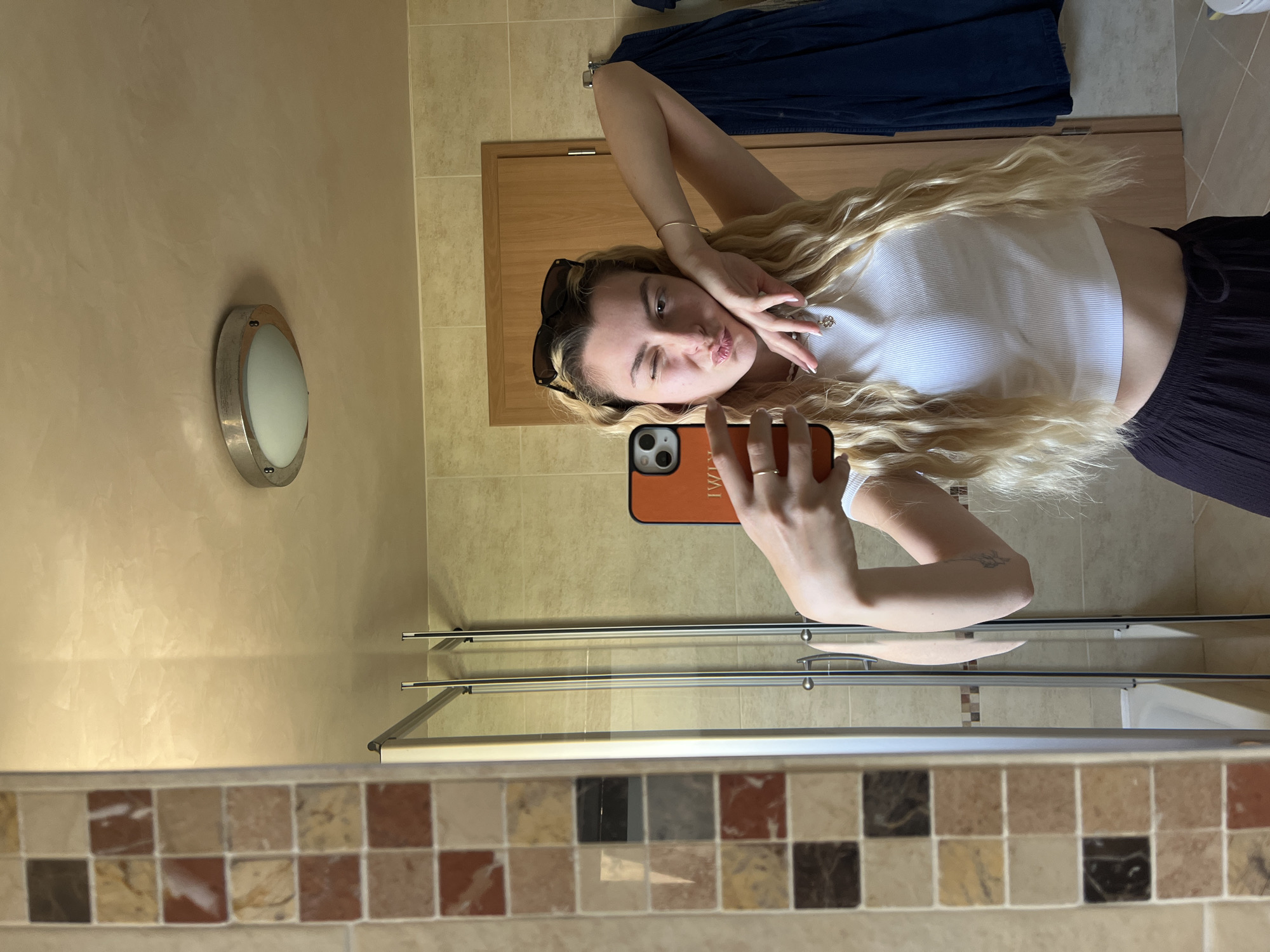 A playful mirror selfie captures a young woman with long, wavy blonde hair. She is wearing a white ribbed crop top and dark, loose-fitting pants, with sunglasses perched on her head. She strikes a pose, winking and puckering her lips, while holding a smartphone with an orange-brown case in her right hand to take the picture. Her left hand is raised, forming a peace sign gesture.
The setting appears to be a bathroom, as suggested by the mirror, a light beige/tan wall with a round, silver-rimmed light fixture, and a metal towel rack visible below her. A wooden door or cabinet and a blue towel are seen in the upper part of the reflection. The bottom edge of the mirror's frame features a decorative band of small, multi-colored square tiles in shades of beige, brown, and reddish-brown. The floor is tiled with light-colored square tiles. The word "TWIN" is visible in reverse on her phone case, indicating the reflection. The image is oriented sideways, with the woman appearing to lie horizontally across the frame.