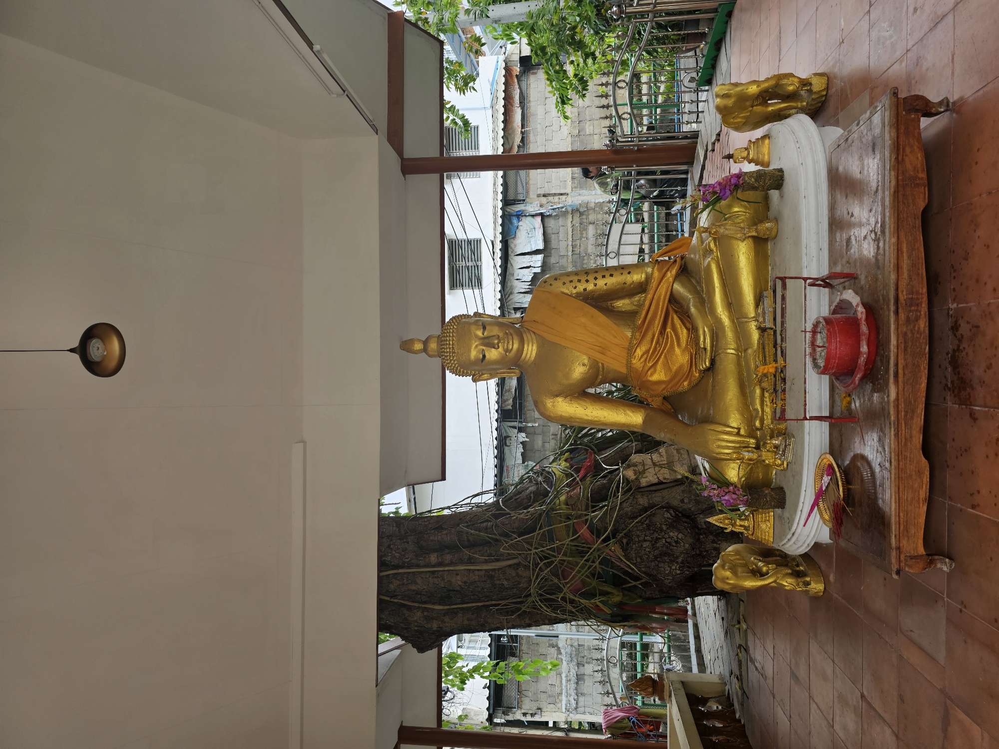 This is a wide-angle, eye-level shot of a golden Buddha statue seated outdoors under a covered pavilion in Bangkok, Thailand. The statue is adorned with a saffron robe and sits on a white, circular platform. To the left and right of the platform are two golden elephant statues, one larger and one smaller. In front of the platform is a low, wooden table with a red incense holder containing lit incense sticks and a brass tray with unlit incense sticks. A large, ancient tree with thick roots and colorful ribbons tied around it is positioned behind the Buddha statue to the left. In the background, behind a metal railing, there are several buildings, some appearing to be residential, with visible electrical wires and foliage. A person wearing a green jacket is partially visible in the background near the railing. The ground is tiled with reddish-brown square tiles. A modern light fixture hangs from the pavilion ceiling.