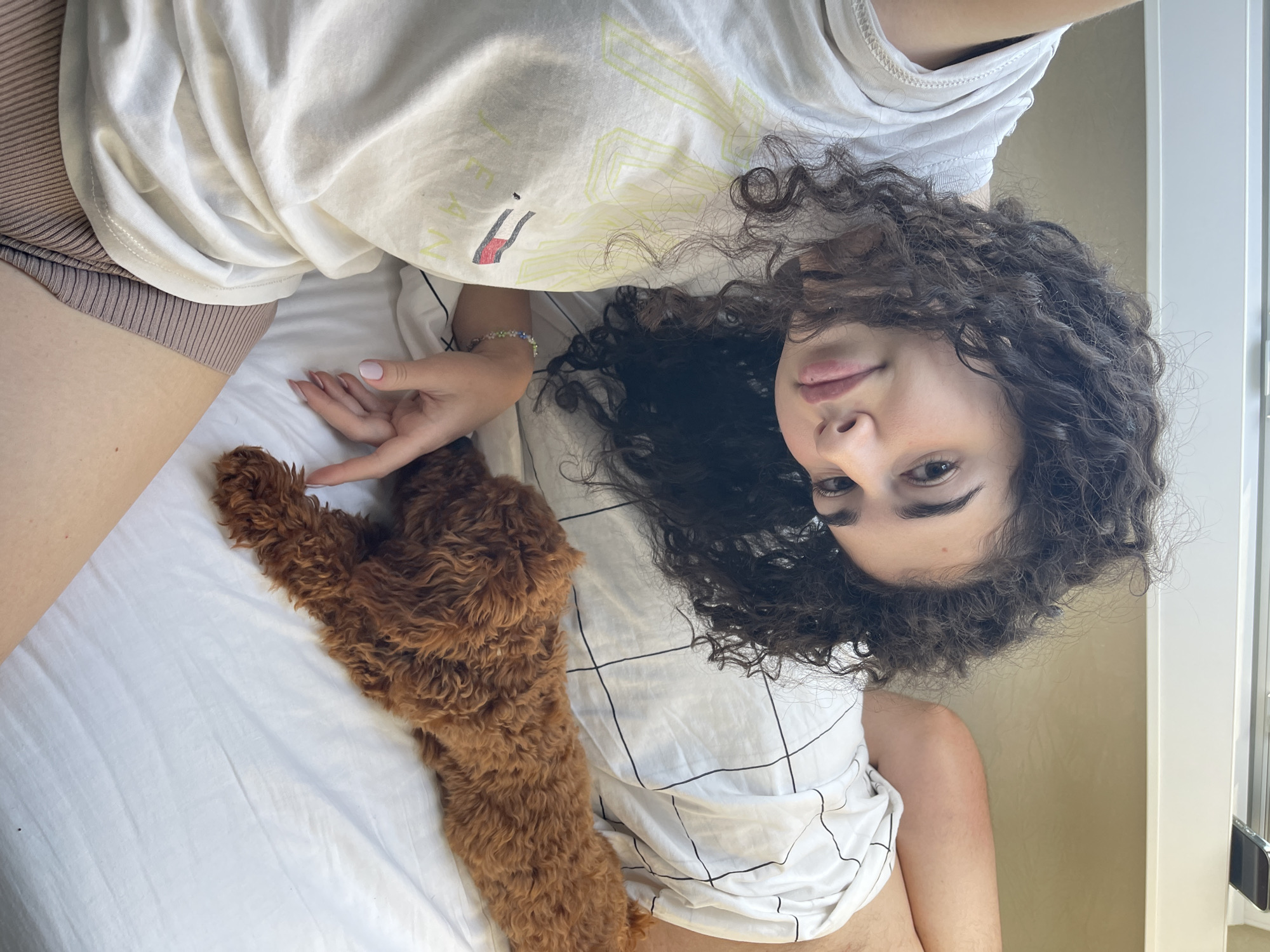 The image captures a young woman and a small dog lying on a bed in Koprinka, Bulgaria. The woman, positioned on her left side, has dark, curly hair and is looking directly at the camera. She is wearing a light-colored t-shirt with a faded yellow "TOMMY JEANS" logo on the front and ribbed brown shorts. Her left arm is visible, adorned with a multi-colored beaded bracelet, and her fingernails display light pink polish. To her right, a small, reddish-brown dog with curly fur lies on its stomach, facing away from the camera. The bedding consists of white sheets and a pillow with a subtle black grid pattern. In the background, a white headboard is visible, with a portion of a black smartphone resting on it. A light beige wall is present above the headboard. The scene is illuminated by natural light.
