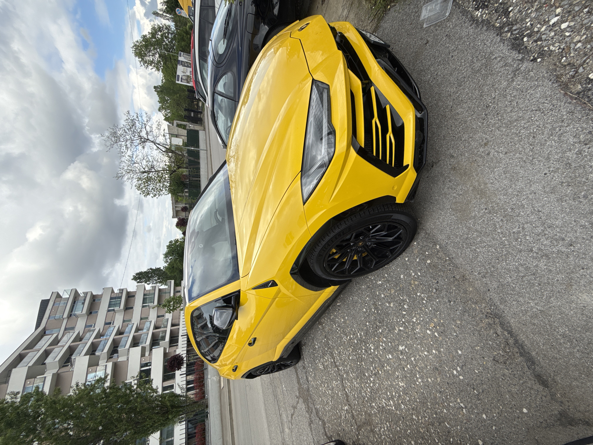 The image shows a yellow Lamborghini Urus parked on a paved street. A dark-colored SUV is parked next to it. In the background, there is a modern apartment building with a unique, staggered facade, along with trees and a cloudy sky. This image was likely taken in Sofia, Bulgaria.