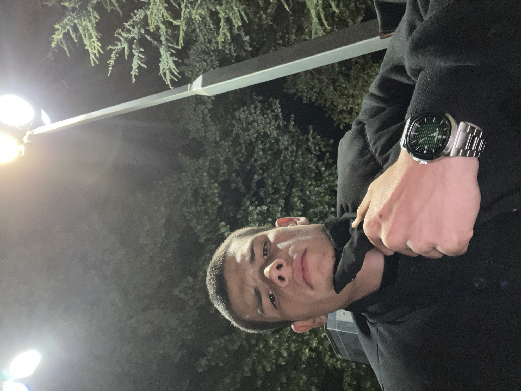 A man is standing outside at night. He is wearing a dark jacket. He has a watch on his left wrist. He is holding the collar of his jacket up to his neck. There are lights behind him and trees in the background. The location is Karavelovo, Bulgaria.