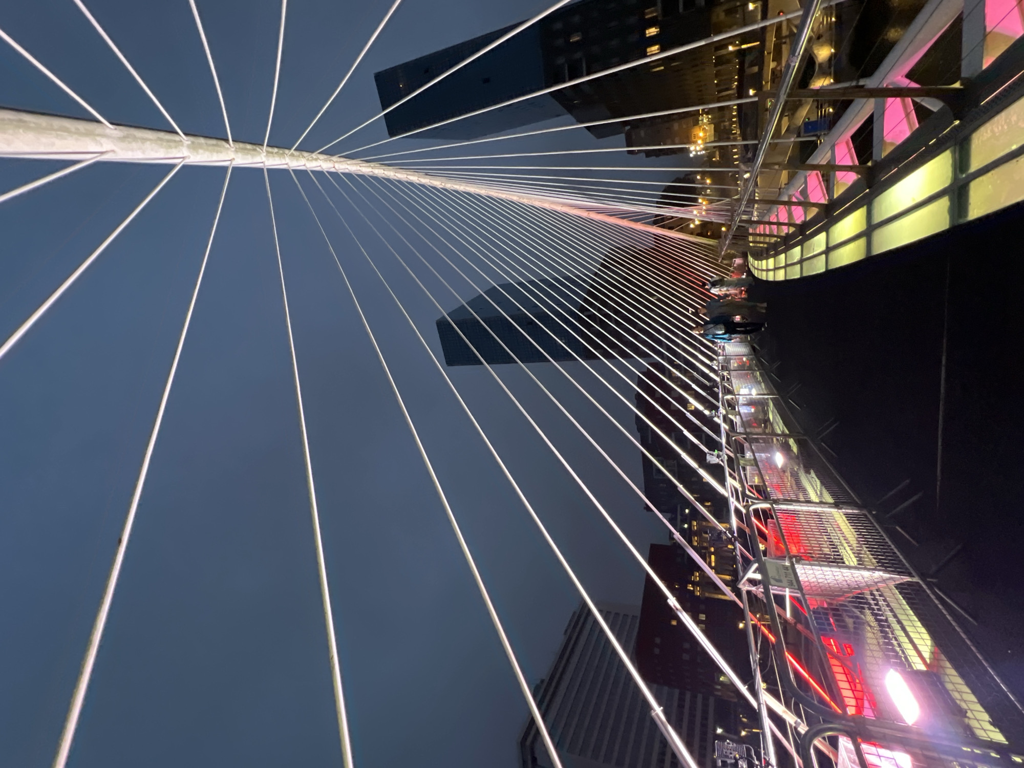 The image depicts the Zubizuri Bridge in Bilbao, Spain, at dusk. The pedestrian bridge, designed by Santiago Calatrava, features a striking white cable-stayed structure that fans out against a darkening sky. Numerous taut white cables extend from a central white mast, creating a geometric pattern. The walkway of the bridge is illuminated with a vibrant display of colored lights, transitioning from pink to yellow, casting a glow on the surface.
Several pedestrians are visible walking along the bridge, appearing as small figures against the grand architectural elements. On the left side of the bridge, red lights from passing vehicles create streaks, suggesting traffic on a nearby road or waterway. In the background, modern glass-fronted skyscrapers with illuminated windows rise, characteristic of Bilbao's contemporary urban landscape. The overall scene captures the bridge's illumination and its integration into the city's nighttime environment.