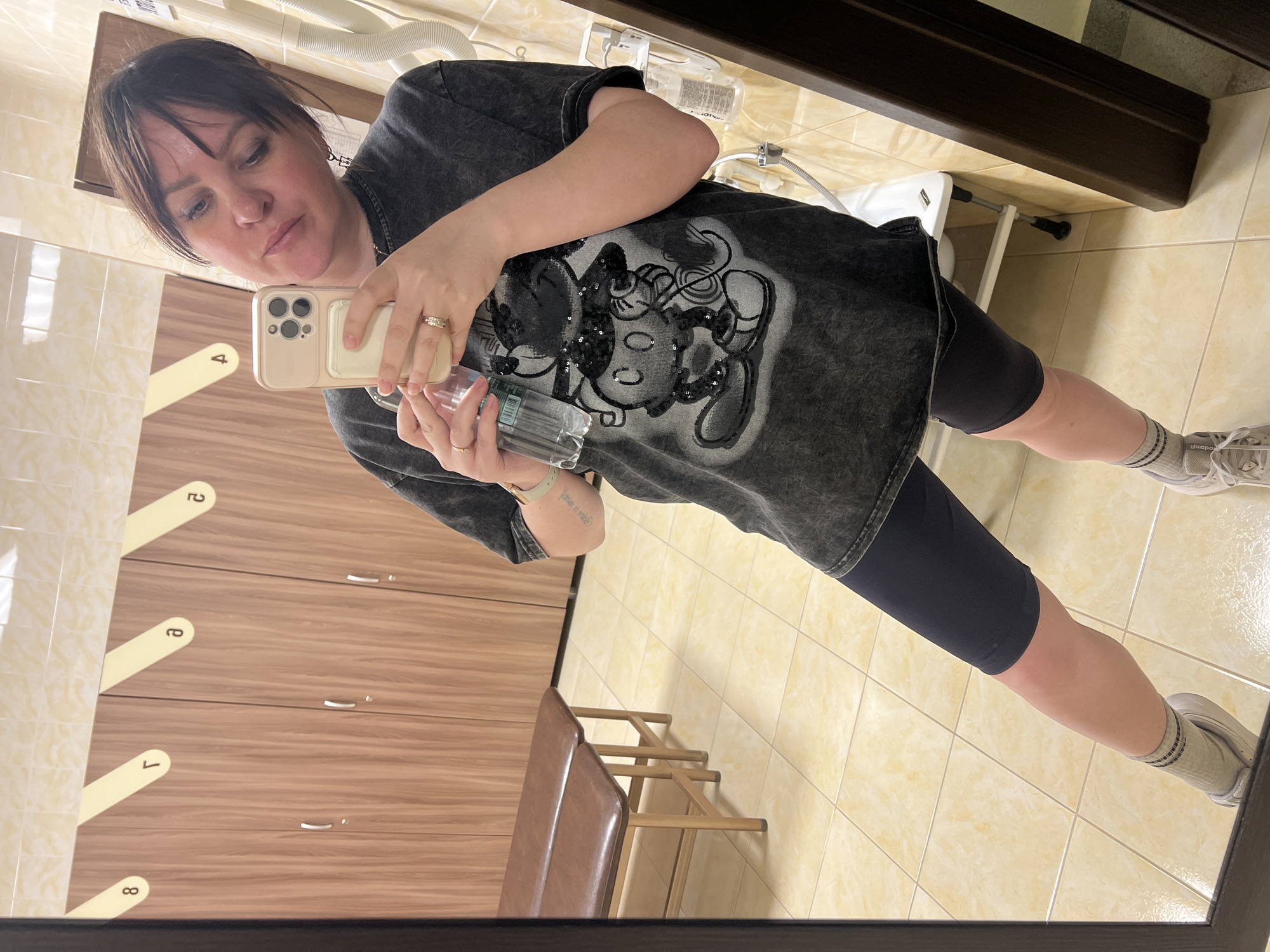 A female individual is depicted taking a mirror selfie in what appears to be a changing room or locker facility. The individual, with dark hair and visible makeup, is wearing a distressed dark grey t-shirt featuring a sequined Mickey Mouse design, black cycling shorts, grey striped ankle socks, and white athletic shoes. She wears a gold-colored ring on her left ring finger and pinky, and a light-colored watch on her left wrist; a small tattoo is present on her left forearm. Her right hand holds a smartphone in a light-colored case, pointed towards herself, while her left hand holds a clear plastic water bottle.
The background shows a row of light brown wooden lockers with silver-colored handles and keyholes. Yellow, angled panels above the lockers display Cyrillic numerals/letters (e.g., '8', 'Г', 'Թ', '೭'). To the left, two long, upholstered brown benches with light-colored frames are situated in front of the lockers. The floor and lower wall are covered in light beige square tiles, while the upper wall features similar, glossier tiles. On the right, a white wall-mounted hair dryer with a corrugated hose, a white specialized fixture (potentially a shower seat or basin) with a faucet, and a wall-mounted dispenser are visible. The specific location in the city of Noviy, Russia, cannot be determined from the visual evidence.