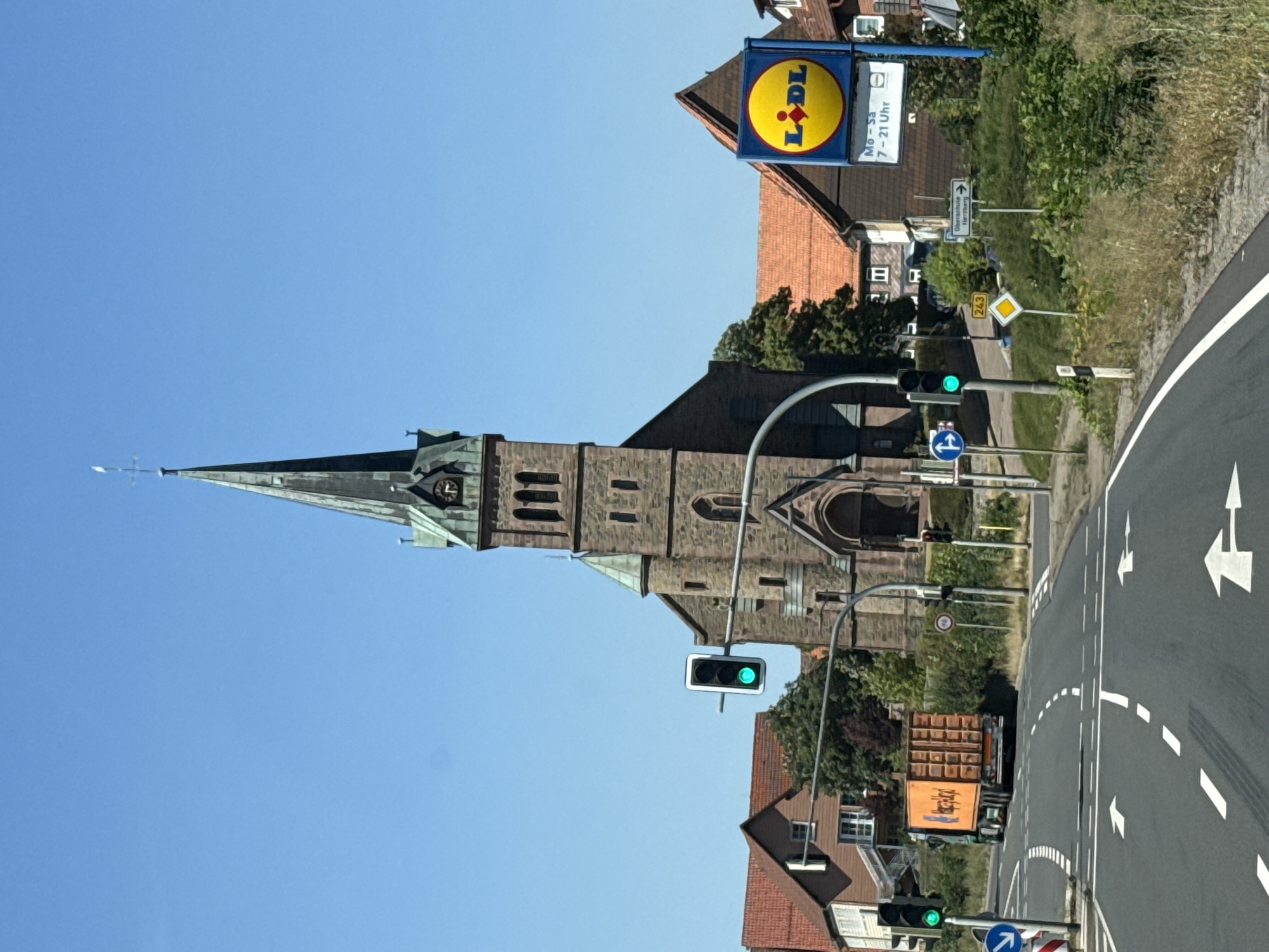 A medium-shot, eye-level, vertical, outdoor, daylight, full-color, photographic image shows a street scene in Herzberg, Germany. The primary subject is a brick church with a tall, pointed steeple that dominates the background. The church has arched windows and a clock near the top of the steeple. To the right of the church, a Lidl grocery store sign is prominently displayed. In the foreground, a two-lane road curves to the left. A traffic light with a green signal is visible on the left side of the road, and another traffic light with a green signal is further down on the right. An orange truck with a trailer is driving away from the viewer on the left lane of the road. Residential buildings with red-tiled roofs are visible on either side of the street, behind the church and the Lidl sign. The sky is clear and blue. A variety of traffic signs and road markings are present, including arrows on the road indicating lane directions and a blue circular sign with a white arrow pointing left. Vegetation, including trees and bushes, is visible along the roadside.