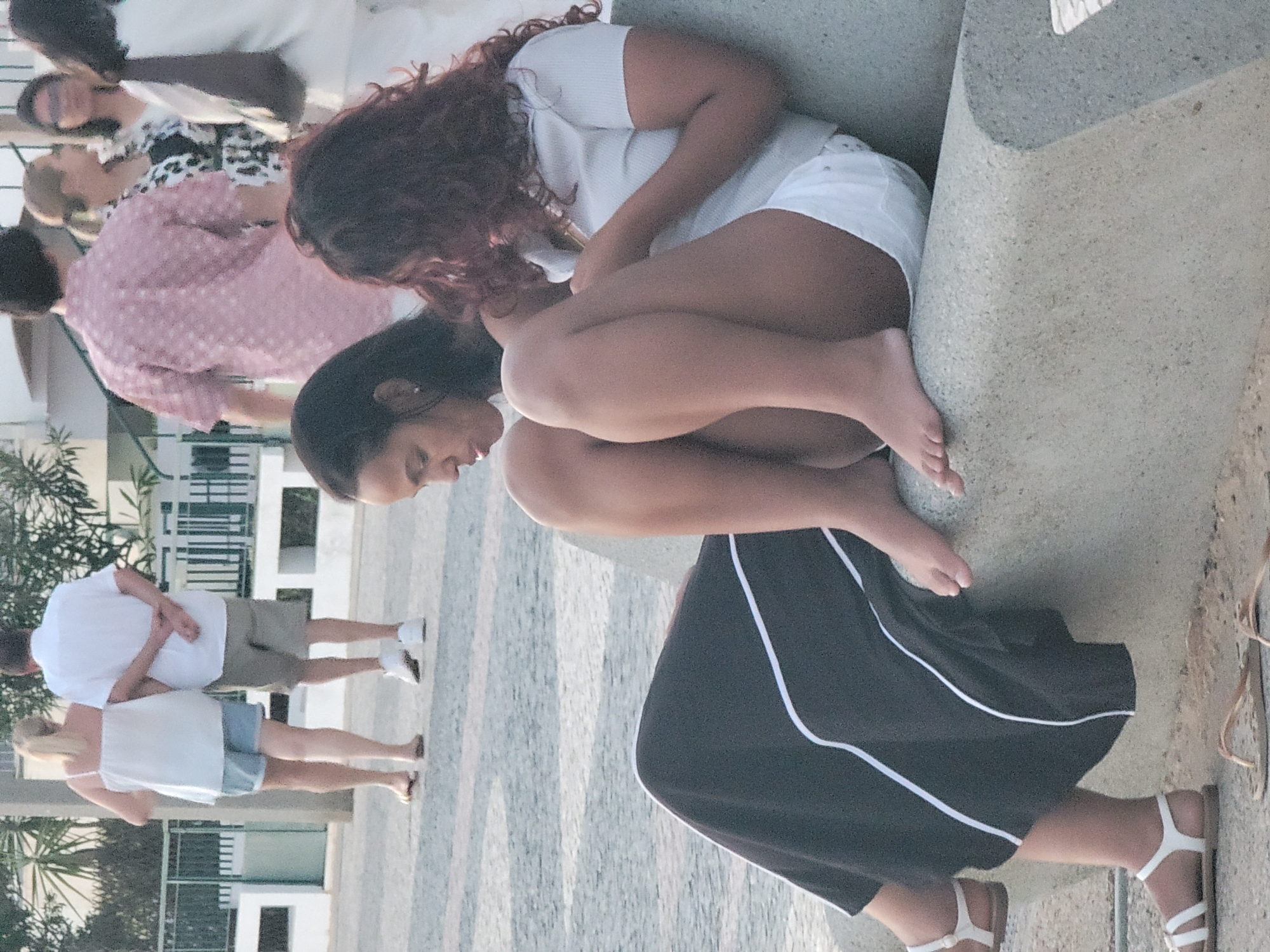 In Quarteira, Portugal, two women are seated on a concrete bench. The woman on the left wears a black skirt with white trim and white sandals. The woman on the right has curly brown hair, wears a light-colored short-sleeved top, and white shorts. She is holding a mobile phone. In the background, several other people are visible, including a man in a pink patterned shirt and a woman with dark hair wearing glasses. The setting appears to be a pedestrian area with a paved ground surface and greenery in the distance.
