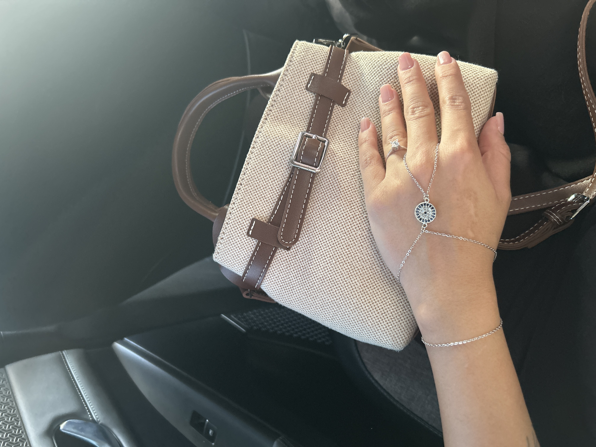 A person's left hand, adorned with a silver bracelet featuring a circular charm and a diamond ring, rests on a light-colored canvas handbag with brown leather accents. The handbag is positioned in the passenger seat of a vehicle. The background shows the dark interior of the car, including a black leather door panel and part of a seat. The scene is captured in bright, natural light, suggesting it is daytime. The image does not provide any specific geographical information placing it in Baku, Azerbaijan.