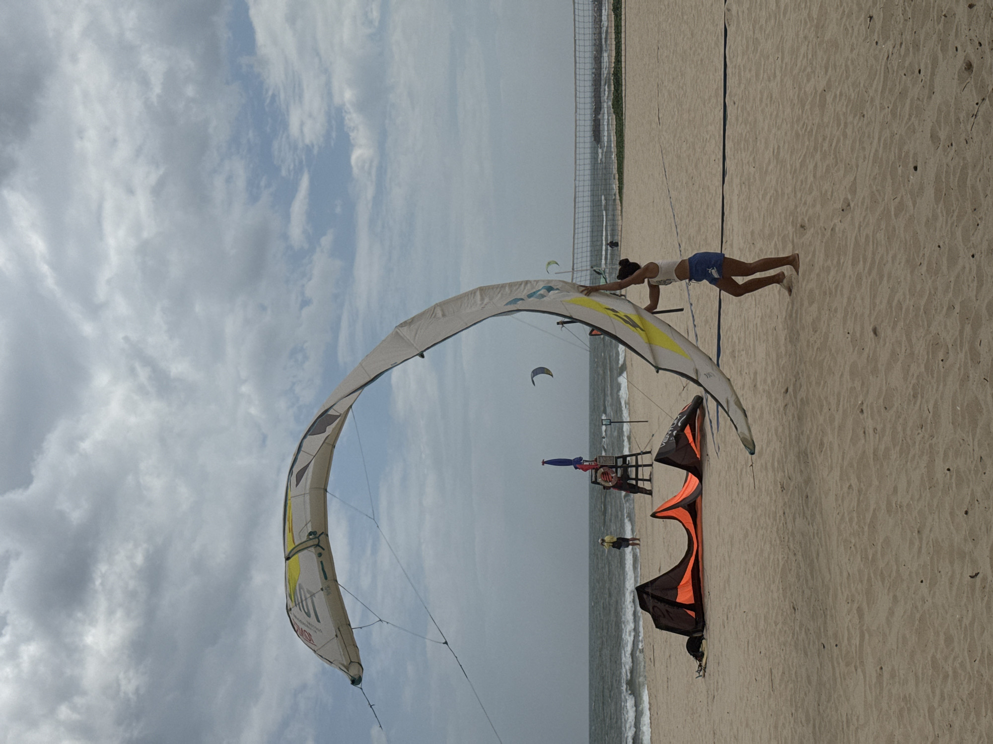 A person stands on a sandy beach in Mũi Né, Vietnam, holding a large kite. The kite is predominantly white with yellow and black accents and is curved upwards. The person is wearing a white tank top and blue shorts. In the background, the ocean is visible with small waves and a cloudy sky. Further back, a volleyball net is set up on the beach. Several other kites are on the sand in the foreground, one being orange and black. A lifeguard chair with flags is also present near the water's edge.