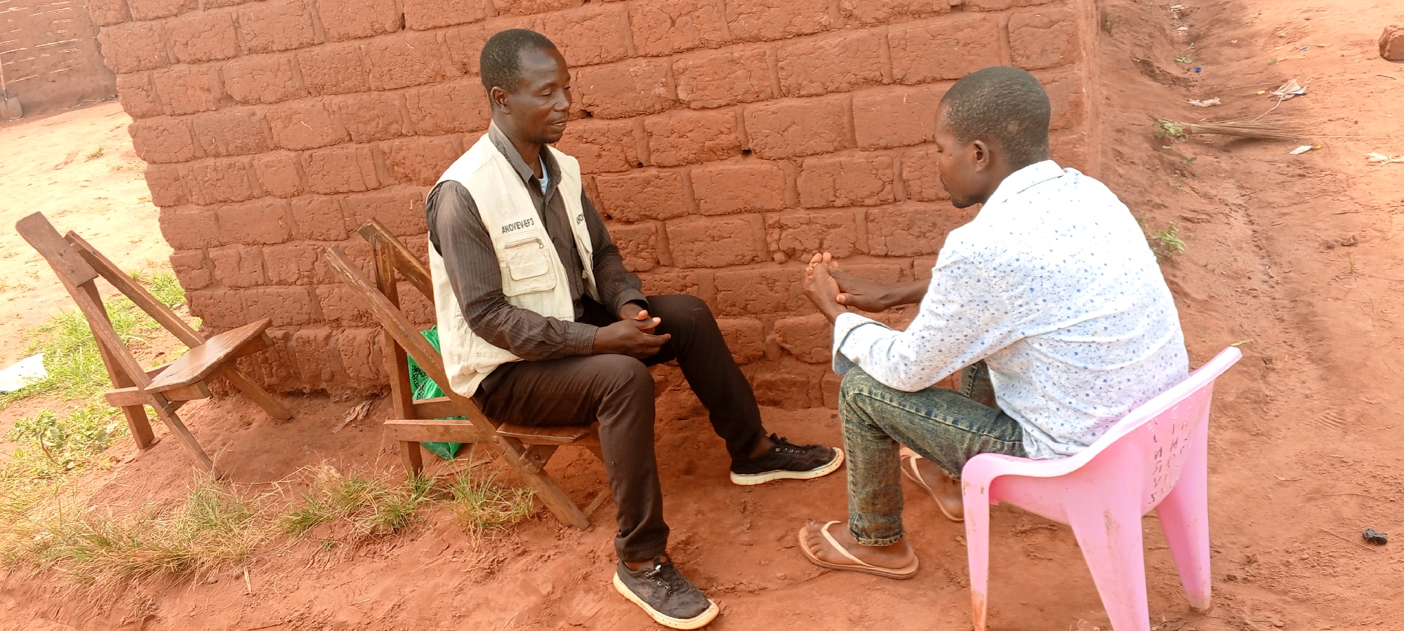 Two dark-skinned men are seated outdoors, engaged in a conversation in Makungu, Democratic Republic of the Congo. The man on the left sits on a wooden folding chair, dressed in a dark long-sleeved shirt, black trousers, and a light-colored utility vest with multiple pockets. Visible text on his vest pockets includes "AMOYEVEF3" and a partial "AND". He looks towards the man on the right with a slight smile.
The man on the right is seated on a pink plastic chair, wearing a light-colored long-sleeved shirt with small blue patterns, blue jeans, and light-colored sandals. He faces the man on the left, with his hands clasped together in front of him. Some faint, unreadable letters are etched or written on the back of his pink chair.
The setting features a building made of reddish-brown mud bricks in the background, indicative of local construction. The ground is composed of reddish-brown earth with scattered patches of green grass. An empty wooden folding chair is visible on the far left. In the distant background to the right, a shallow trench or ditch runs along the ground, containing some dried leaves or debris. The scene is illuminated by natural daylight, suggesting it is daytime, possibly morning or afternoon, with soft, diffused light and no harsh shadows.