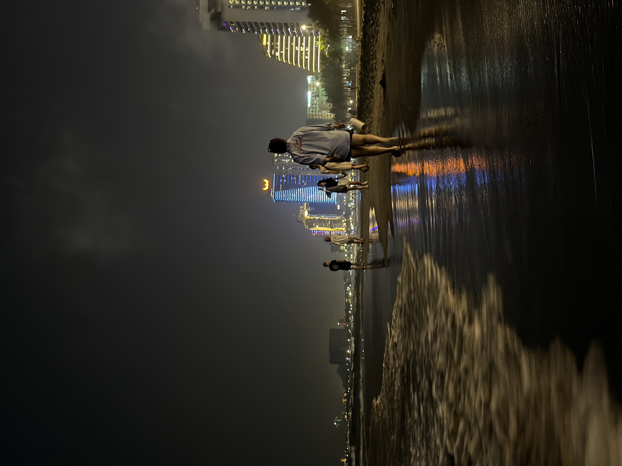 At night, a group of people stroll along a beach in Đà Nẵng, Vietnam. The wet sand reflects the city lights, blurring the boundary between the beach and the city skyline. The backdrop shows illuminated skyscrapers and buildings. The foreground features a sandy beach being met with the ocean. There are indistinct buildings in the background. The sky is dark, and the focus is on the illuminated structures along the coast and the reflections on the water. The primary focus is on the people at the beach and the lights from the city reflecting off the wet sand.