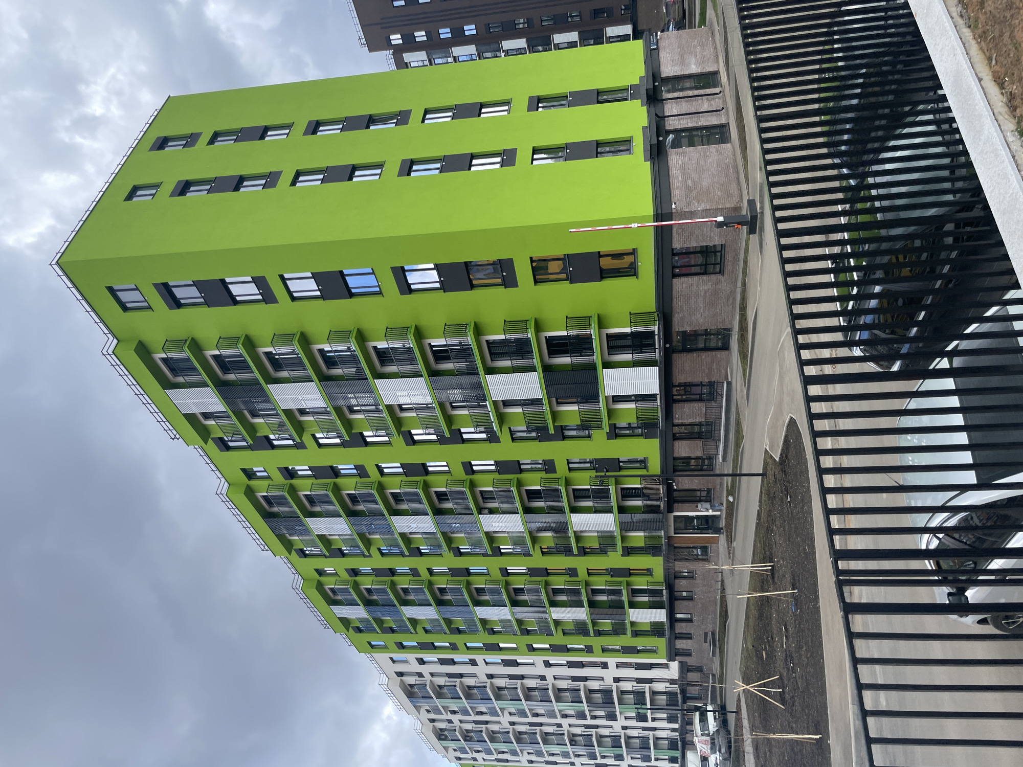 The image captures a modern residential or commercial complex in Pervomaysky, Russia, under an overcast sky. The focal point is a multi-story building painted in a striking lime green, featuring numerous rectangular windows with dark frames and integrated balconies or air conditioning unit covers, some white and some dark. This building exhibits contemporary architectural design with varied protrusions and recessed sections on its facade.
To the right and behind the green building, a taller, dark brown or grey building with multiple stories and windows is visible. Below the green structure, another building with a lighter, white or grey facade extends into the background.
In the mid-ground, a black metal fence with horizontal bars runs along the right side, partially obscuring a paved area with several parked cars. A red and white barrier arm, indicative of a controlled access point, is positioned near the base of the green building. The ground level features patches of bare earth, some with small, staked saplings, and paved pathways. The sky is uniformly cloudy and grey, suggesting an overcast day. No specific human activity is visible, and no text is discernible in the image.