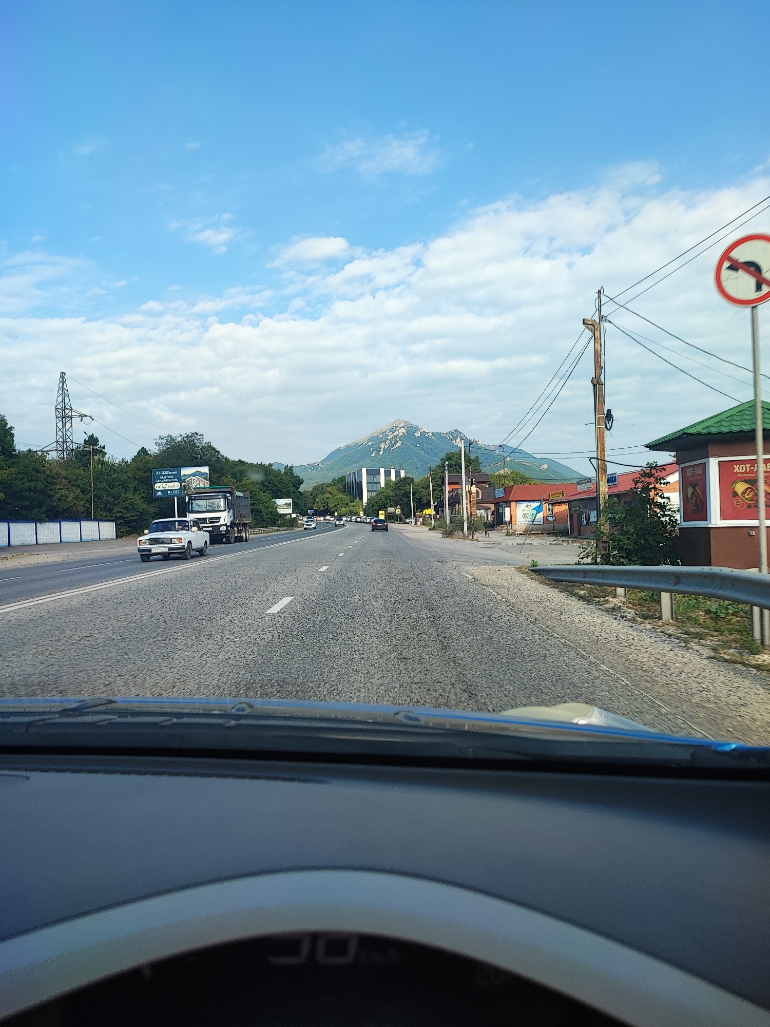 The image shows a view from a car driving down a highway towards a mountainous landscape, likely near the city of Inozemtsevo, Russia. The road is paved and has lane markings. On the left side of the road, there is a white concrete barrier and trees. On the right side, there are buildings, including a building with a green roof, and a utility pole with power lines. A sign prohibiting left turns is visible near the utility pole. Several vehicles, including cars and a truck, are on the road. In the background, a large mountain peak is visible, with some buildings at its base. The sky is blue with scattered white clouds. The front of the car's dashboard and windshield are visible in the foreground.