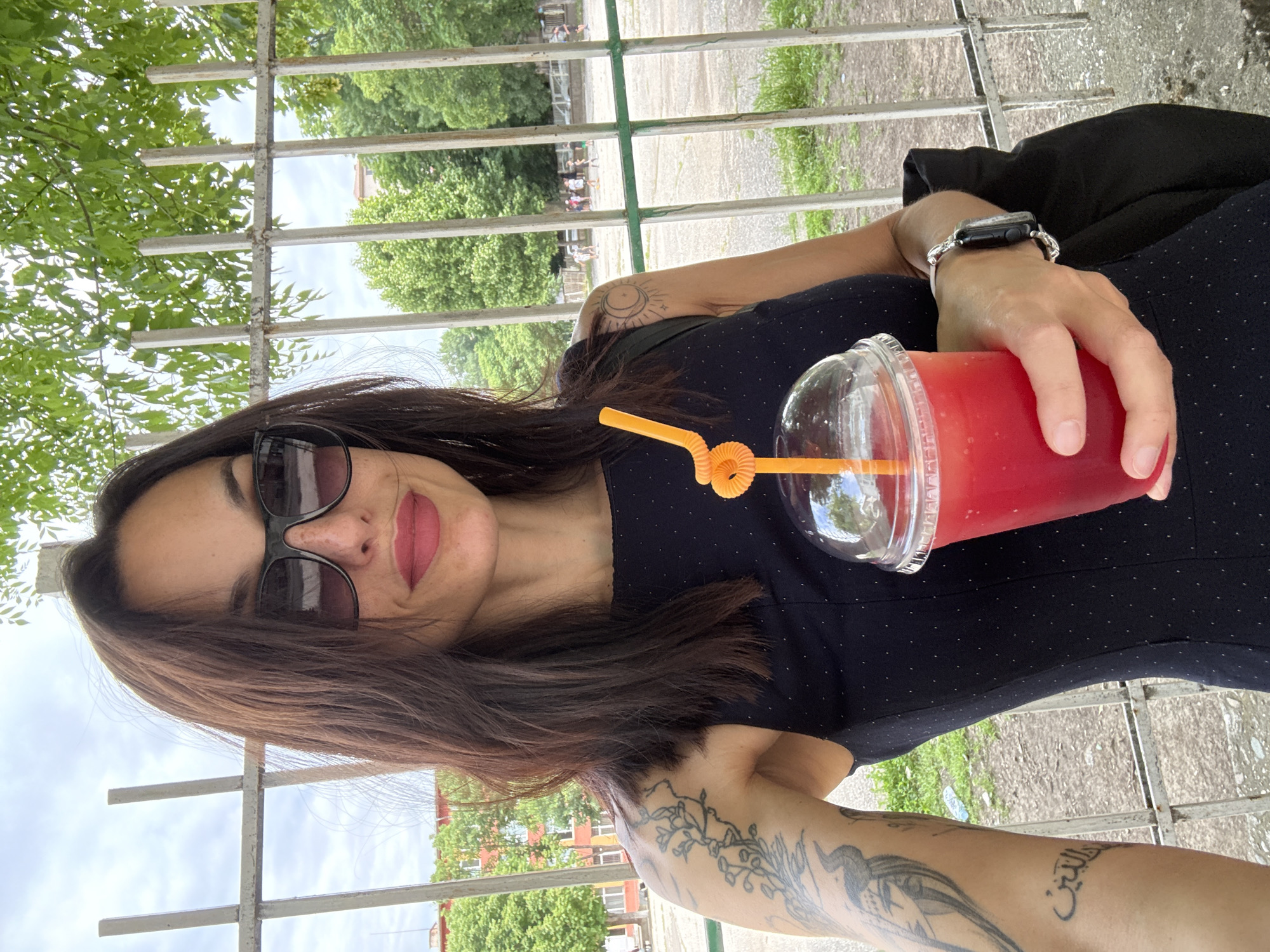 A woman in a dark dress and sunglasses is holding a clear plastic cup with a bright red liquid and an orange straw. She is wearing a smartwatch on her left wrist and has tattoos on both arms. The background shows a metal fence, trees, and a building. Based on the trees and the type of fence, the location is likely in a park or public area. Without further information, precise identification of a location in Pleven, Bulgaria, is not possible.