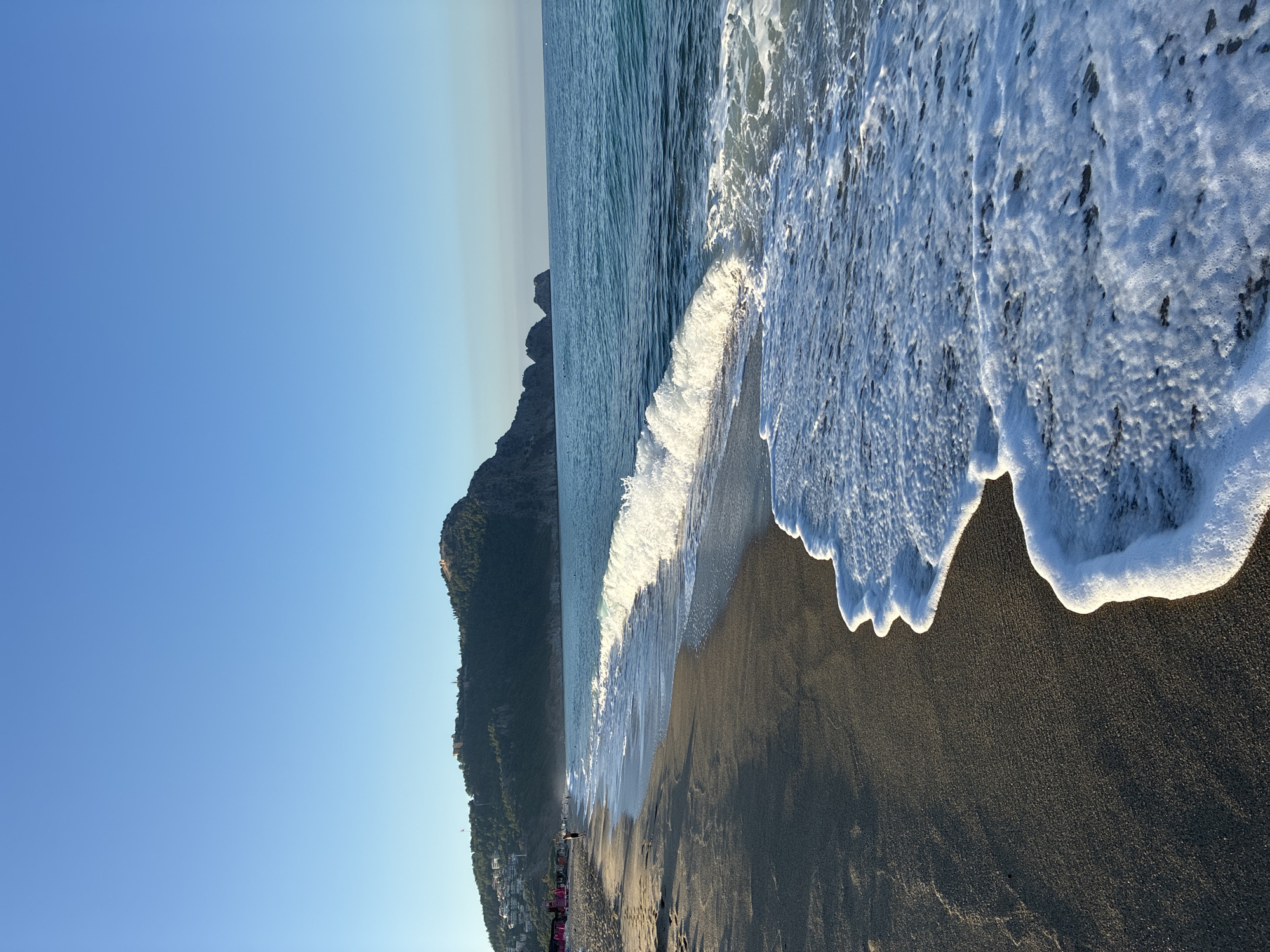 The image captures a serene beach scene in Alanya, Turkey, with the majestic Alanya Castle perched atop a hill in the background. The foreground is dominated by the dark, sandy shore where gentle waves of the Mediterranean Sea are rolling in. The water is a clear, inviting blue, and the sunlight glints off the breaking waves, creating a foamy white crest.
Further down the beach, a few beach chairs with pink umbrellas are visible, suggesting a leisurely atmosphere. A lone figure stands near the water's edge, facing the sea, adding a sense of human presence to the vastness of the landscape. The hill leading up to the castle is covered in lush green trees, contrasting with the ancient stone structure that crowns it. The sky is a clear, brilliant blue, typical of a sunny day in this coastal city. The overall impression is one of tranquility and natural beauty, inviting viewers to imagine a peaceful day by the sea in Alanya.