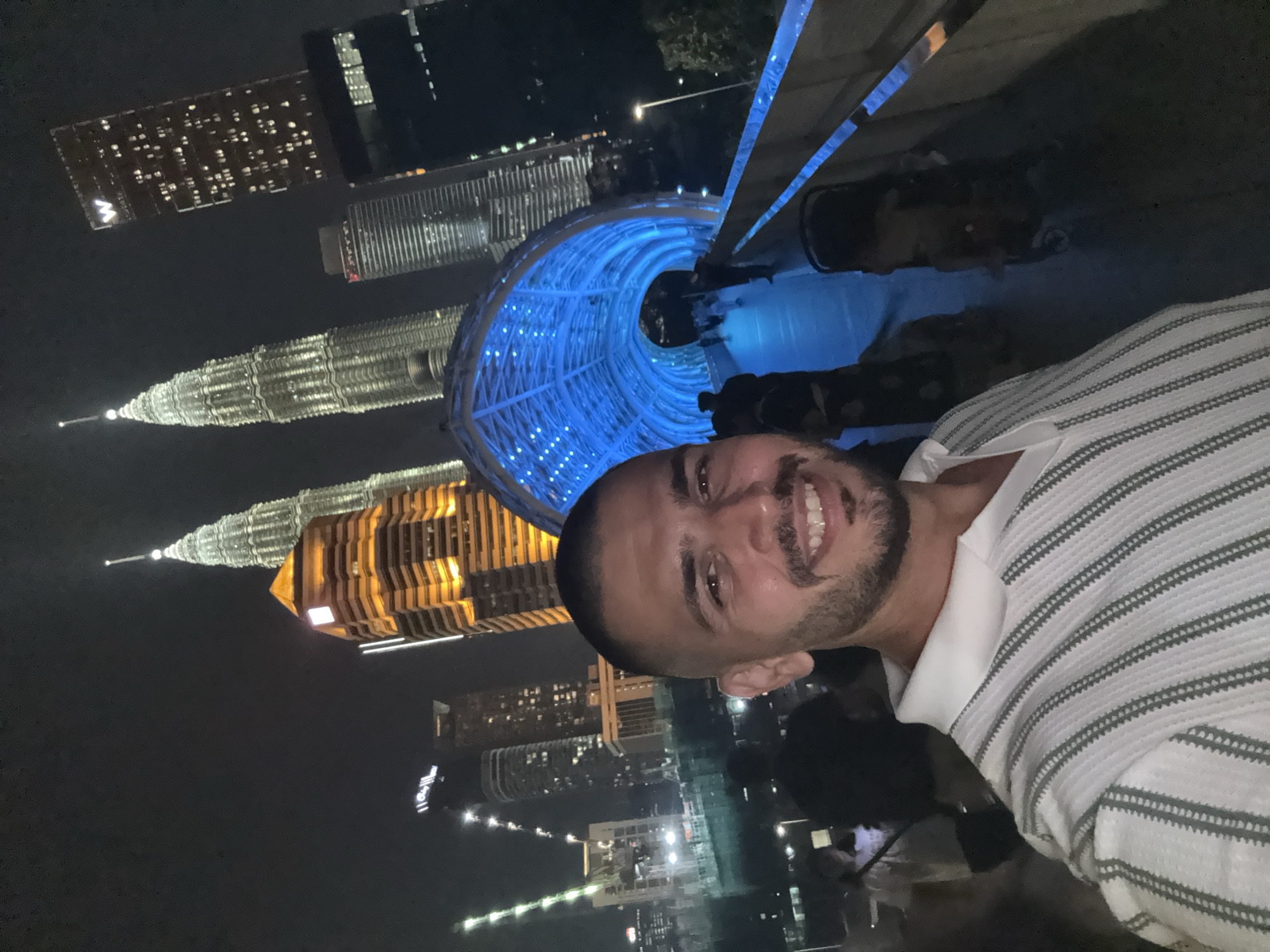 The image shows a man in a white and grey striped shirt taking a selfie at night. Behind him, illuminated in vibrant blue, is a structure resembling a tunnel or archway. In the background are several skyscrapers, including the Petronas Twin Towers, which are lit up in gold. Additional high-rise buildings with illuminated windows are also visible, indicating an urban setting. The scene is likely in Kuala Lumpur, Malaysia, due to the presence of the Petronas Twin Towers. There are also other individuals in the background, further emphasizing a public space at night.