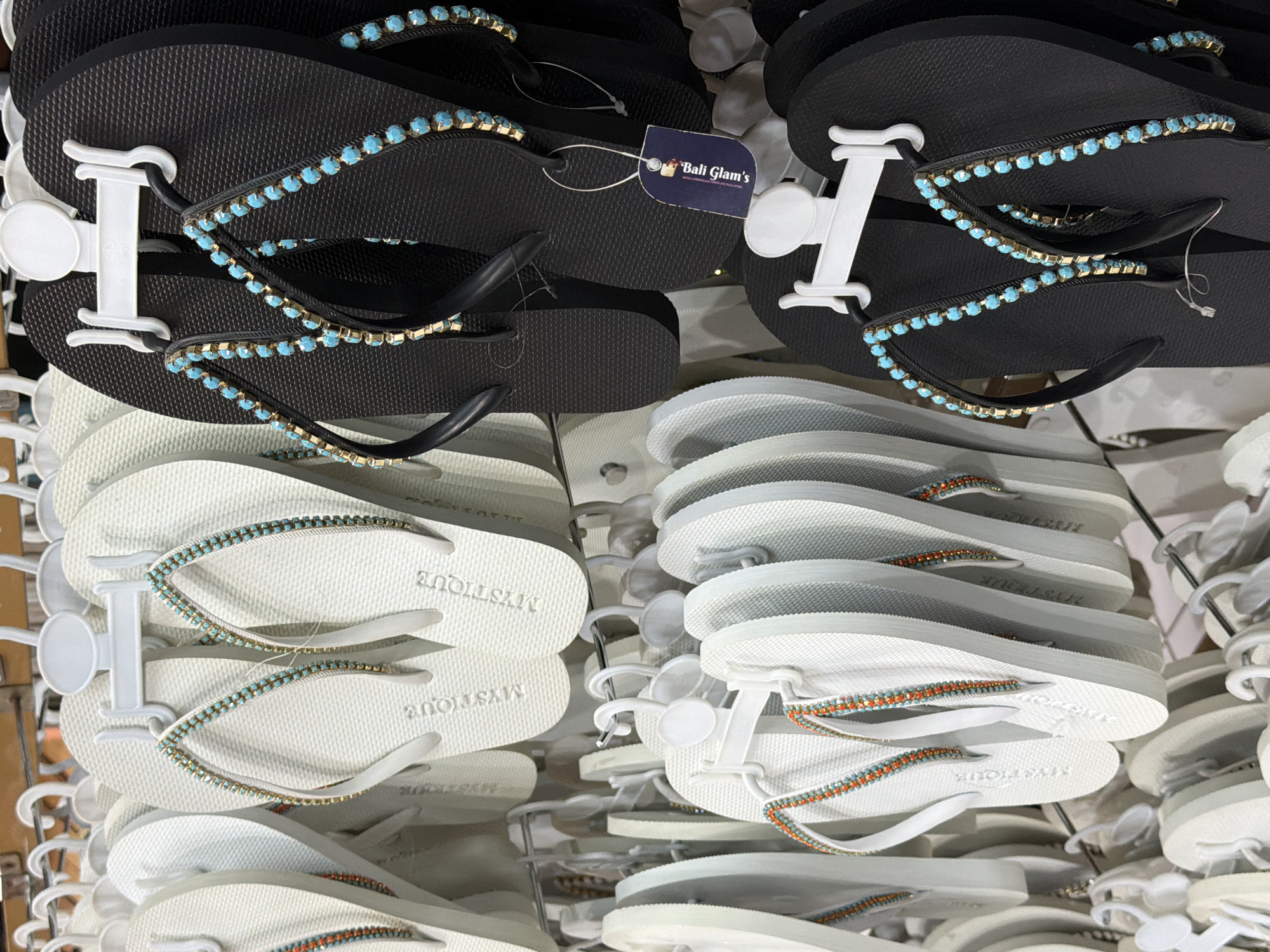 In a retail store in Kuta, Indonesia, a close-up shot captures a display of flip-flops on a hanging rack. The flip-flops are predominantly white and black, with some featuring decorative embellishments on their straps. Specifically, several white flip-flops are adorned with a band of small, alternating turquoise and gold beads. Similarly, a pair of black flip-flops is decorated with turquoise and gold beads. The flip-flops are suspended by white plastic hangers, each clipped onto a metal rod, creating a multi-layered display. The background is blurred, suggesting other merchandise or store fixtures. A small, dark blue tag with the text "Bali Glam's" is attached to one of the black flip-flops. The overall scene depicts a commercial environment focused on selling footwear.