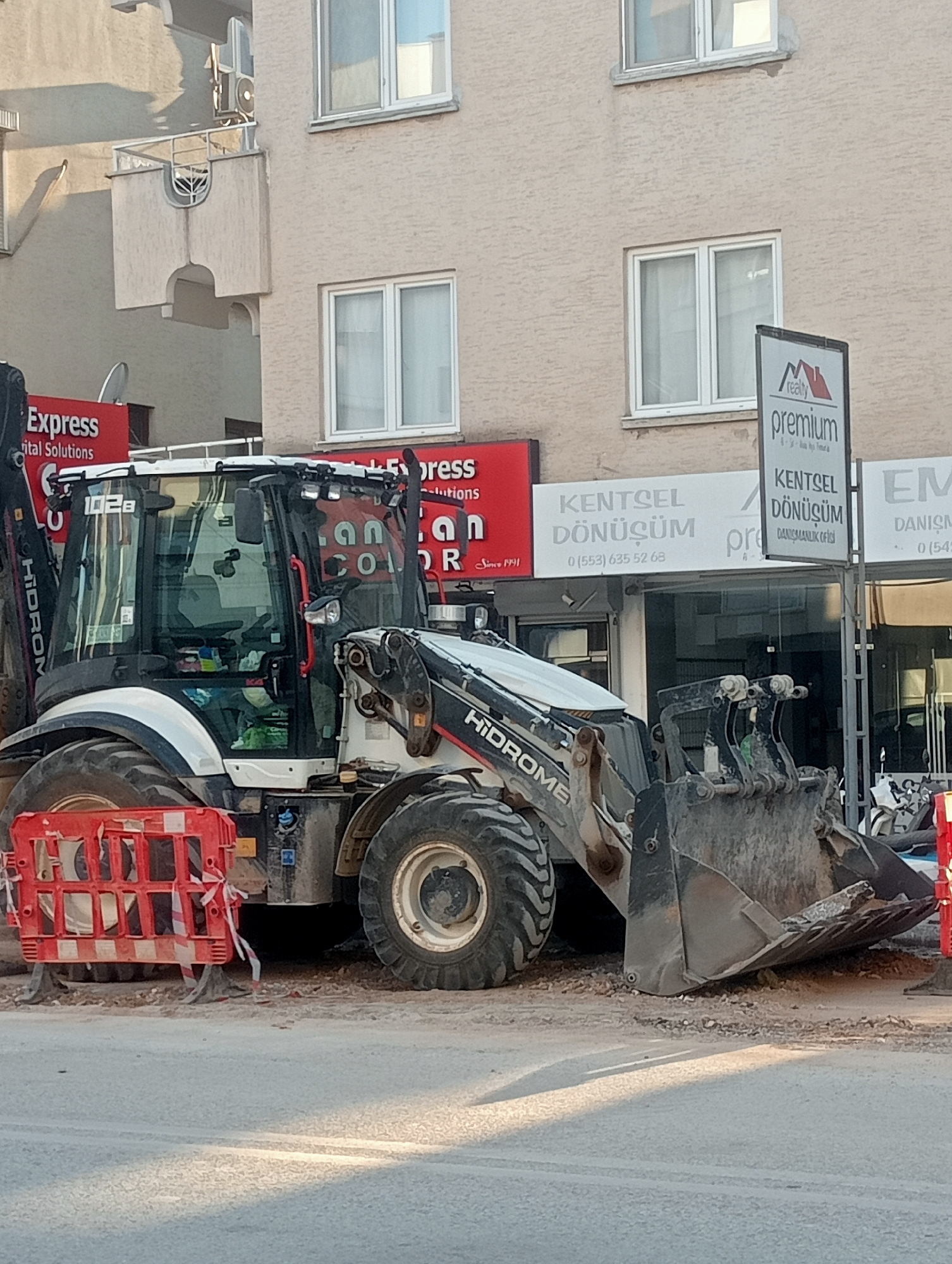An exterior, medium shot captures a construction site in an urban setting. The focal point is a large, white and black backhoe loader, positioned on a dusty, unpaved surface. The vehicle's large tires are prominent, and its bucket is lowered towards the ground. In the background, a light-colored, multi-story building with windows and balconies is visible. Several signs are displayed on the building's facade, one of which reads "premium" in large letters above smaller text. Red construction barriers with white reflective tape are placed in front of the backhoe. The scene is illuminated by natural light, casting shadows on the ground. The overall impression is one of ongoing construction work in a commercial or residential area.