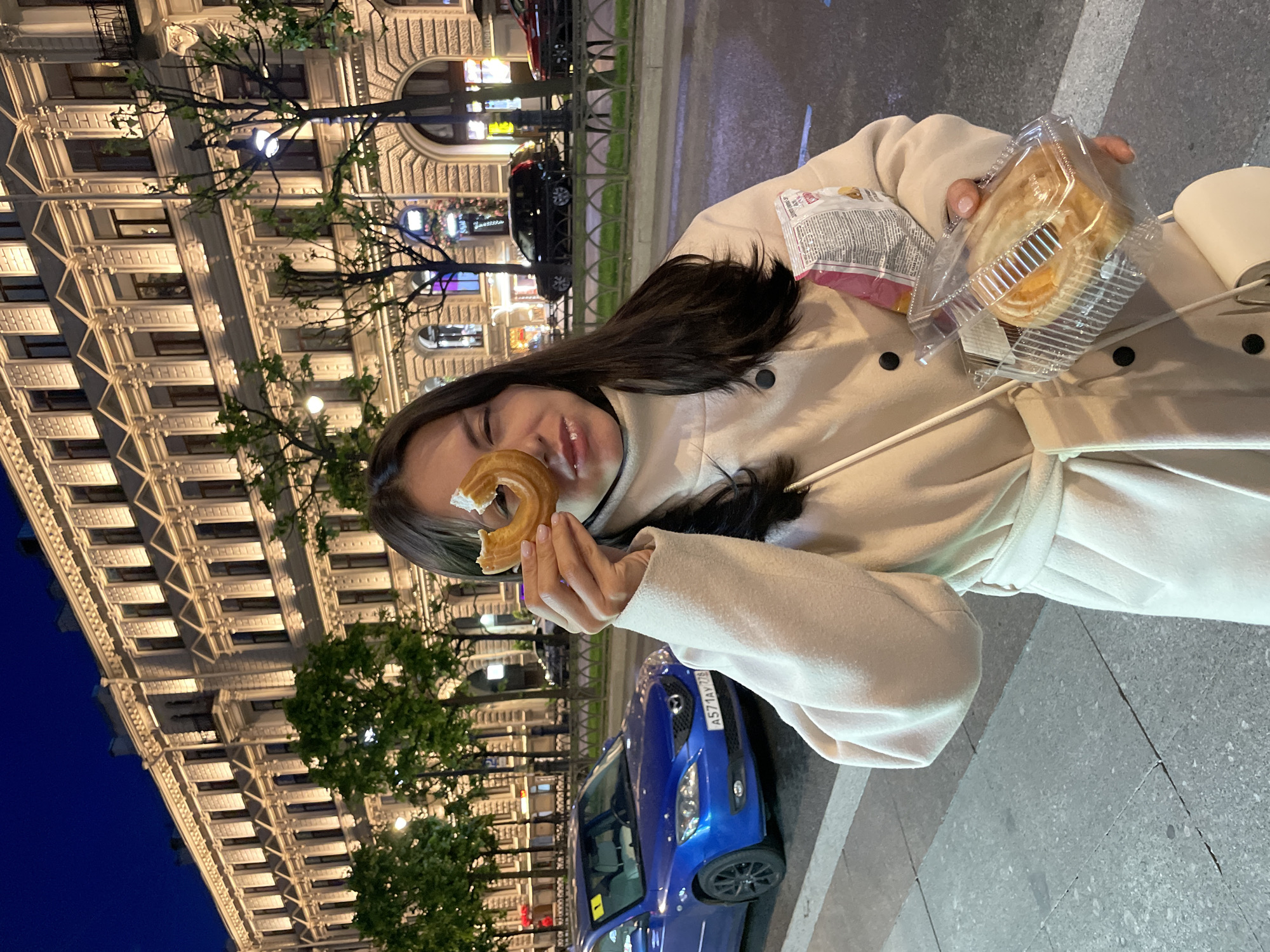 A young woman is standing on a sidewalk at night in front of a building in Saint Petersburg, Russia. She is wearing a cream-colored coat and holding a donut up to her eye. She is also holding a clear plastic container with a pastry inside and a bag of chips. The building behind her is illuminated with lights. A blue car is parked on the street in front of the building.