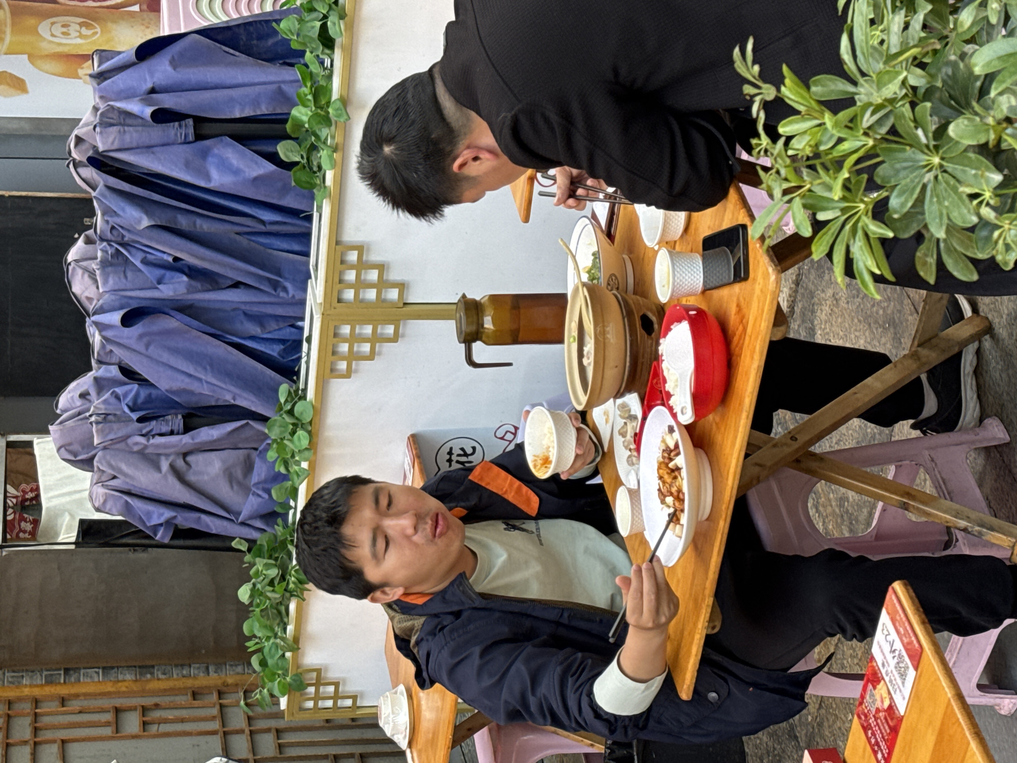 Two men are seated at a wooden table, eating. One man wears a black jacket, and the other wears a blue and orange jacket over a light green t-shirt. They are using chopsticks to eat from bowls and dishes on the table. A brown clay pot sits in the center of the table. The table is set up outdoors, alongside a building with a decorated wall, likely in Jinjiang District, China. Folded purple umbrellas are visible in the background, and there's some greenery as decoration. The location is likely a restaurant or street food stall.