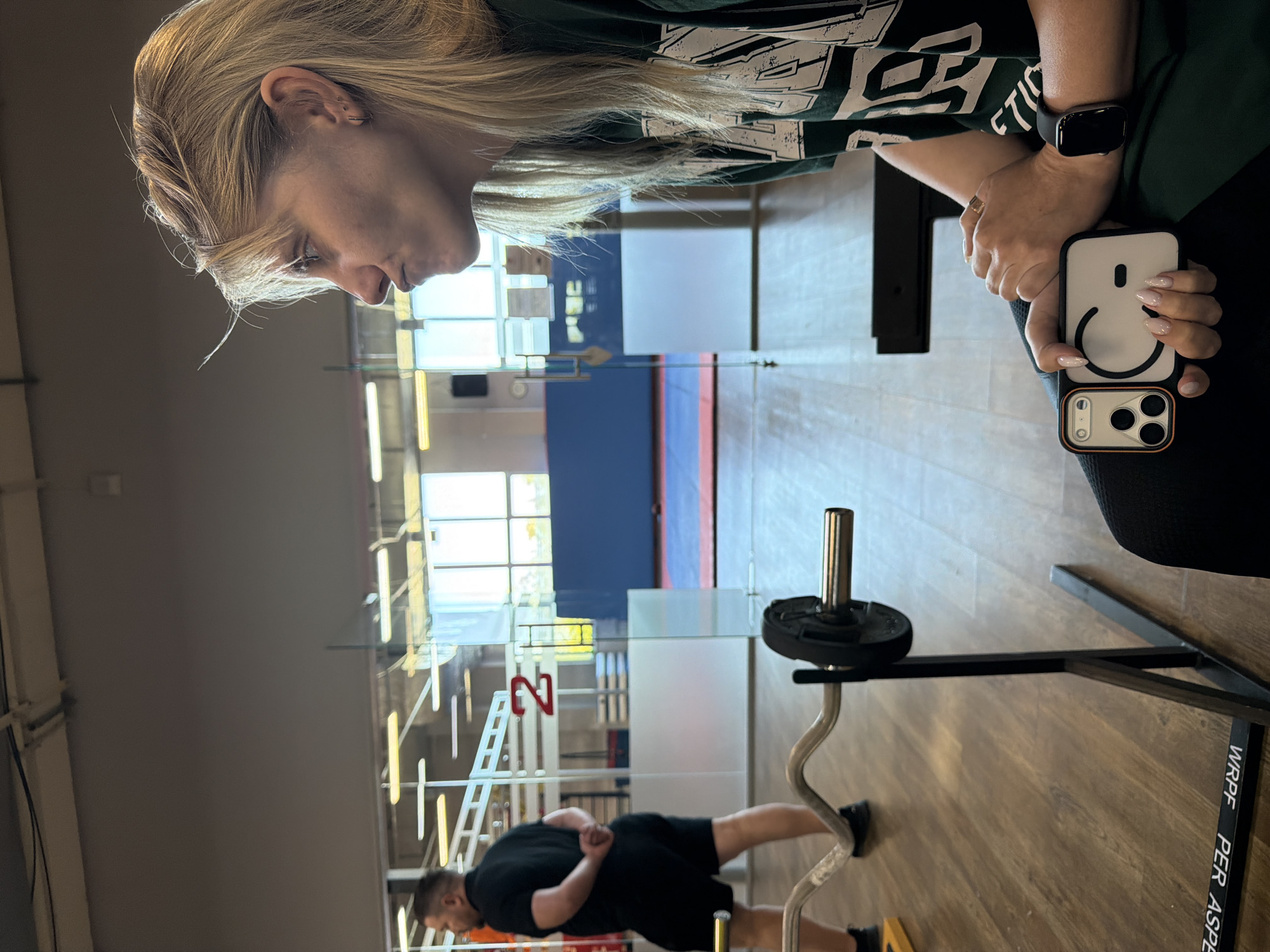 A woman with blonde hair is sitting on a black stool in a gym in Chaikovsky, Russia. She is wearing a green t-shirt and holding a white phone case with a smiley face on it. Her right arm is resting on her lap. She has a ring on her left ring finger and a black smartwatch on her left wrist. In the background, a man in a black t-shirt and black shorts is standing with his hands behind his back. There is a barbell with weights on a rack in the foreground. The gym has wooden floors and large windows that let in natural light.