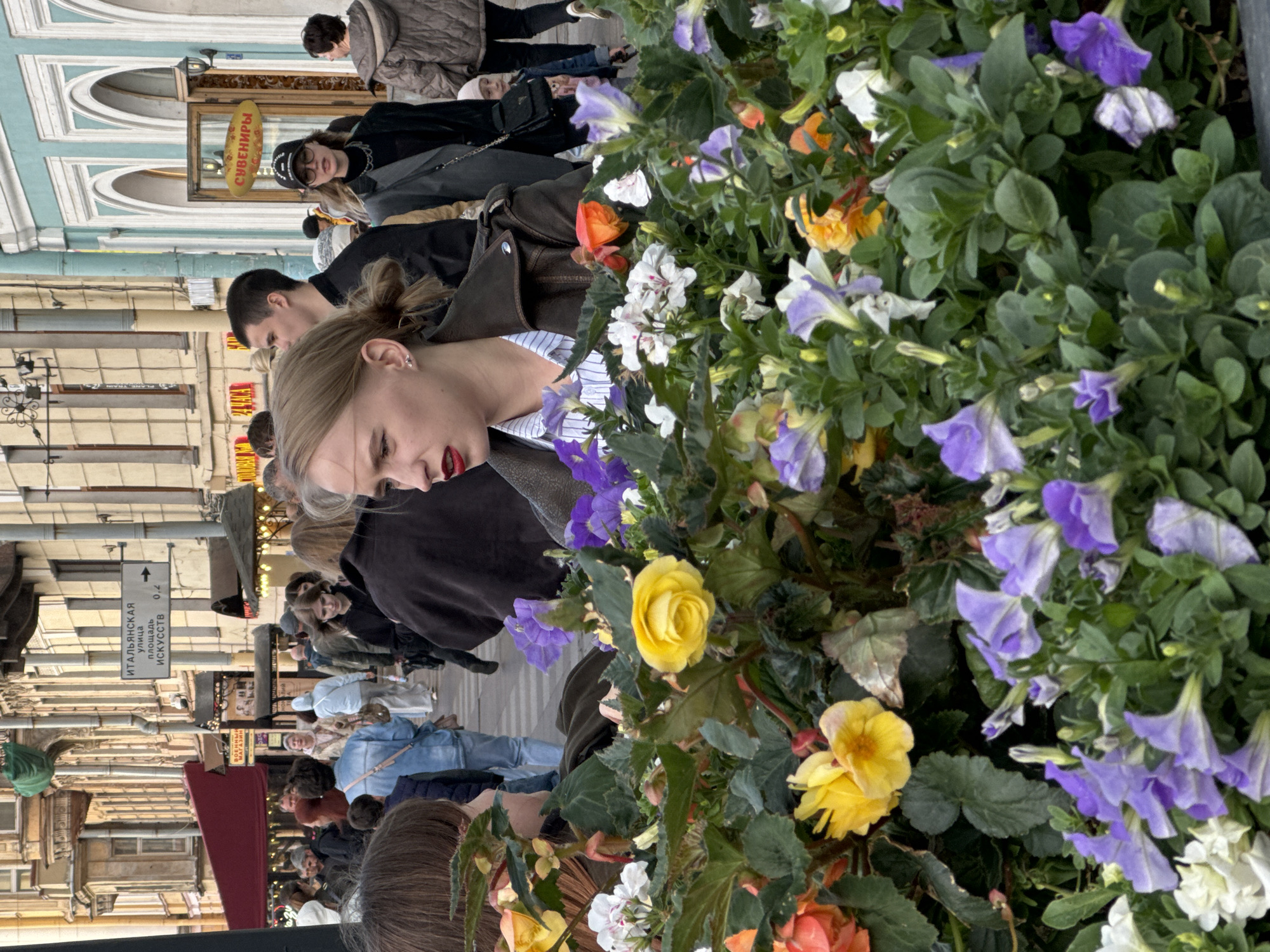 The image depicts a street scene in Saint Petersburg, Russia, likely on Nevsky Prospekt, near the Church of the Savior on Spilled Blood. A variety of people are visible, including women, men, and children. In the foreground, a woman with blonde hair pulled back in a bun and red lipstick is looking to the side. She is wearing a brown leather jacket over a striped shirt. She is in front of a flower bed filled with various colorful flowers, including yellow roses and purple petunias. In the background, people are walking along a sidewalk lined with buildings. Some of the buildings have ornate architecture, and one has a bright blue facade with arched windows. A sign in Russian indicates "Italian Street" and "Square of Arts." The overall atmosphere appears to be that of a busy urban environment, possibly a tourist area.