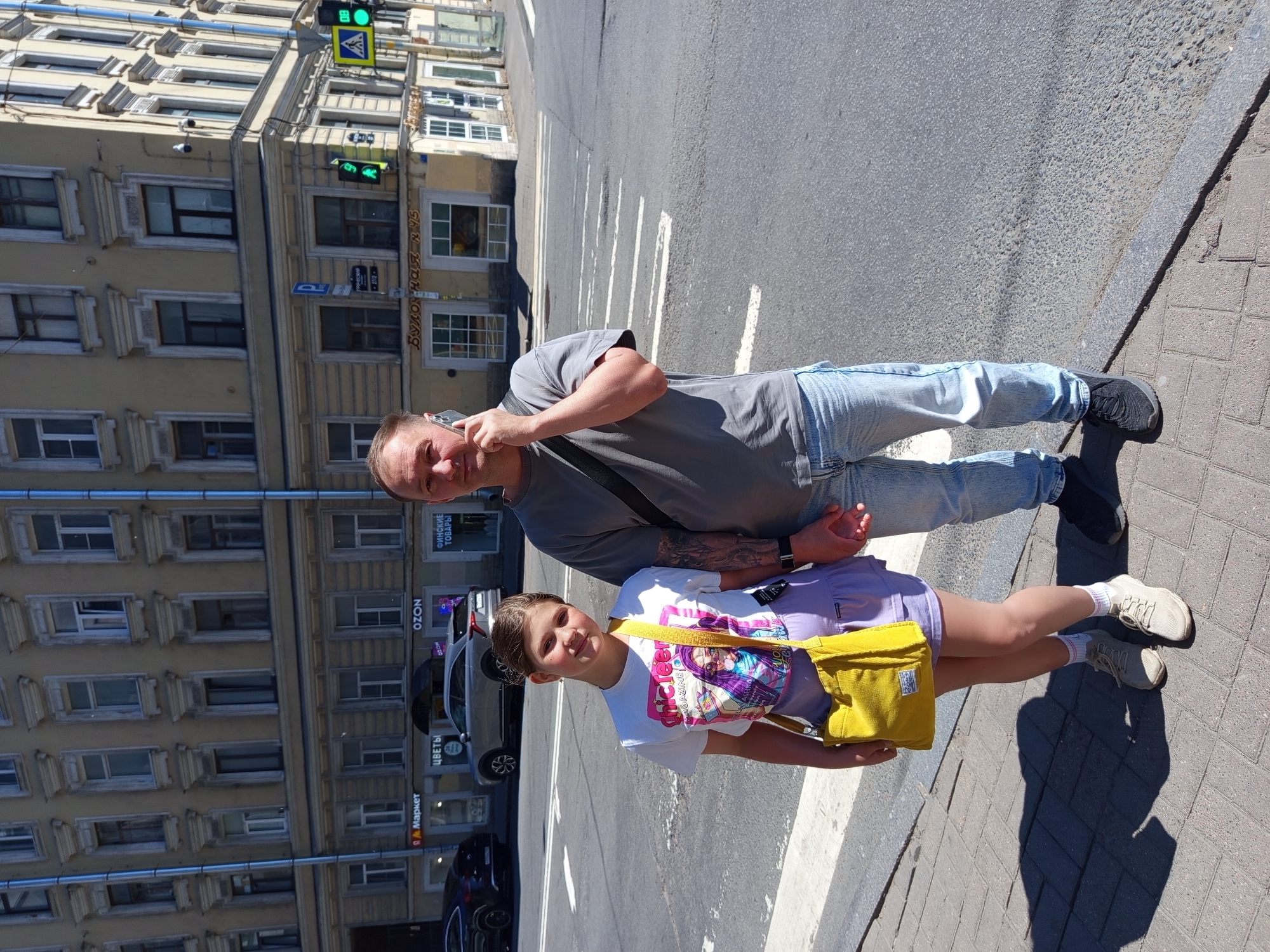 A man and a young girl stand on a sidewalk in front of a building with shops on the ground floor in Saint Petersburg, Russia. The building is light yellow with numerous windows. The man, on the right, is wearing a gray t-shirt, blue jeans, and dark sneakers. He is holding a smartphone to his ear and looking at the camera. The girl, on the left, is wearing a white t-shirt with a graphic print, a light purple skirt, white socks, and light-colored sneakers. She has a yellow shoulder bag. The two are standing close together, with the man's hand lightly touching the girl's hand. The ground is paved with bricks and asphalt. Traffic lights are visible in the background, with a pedestrian crossing marked on the street.