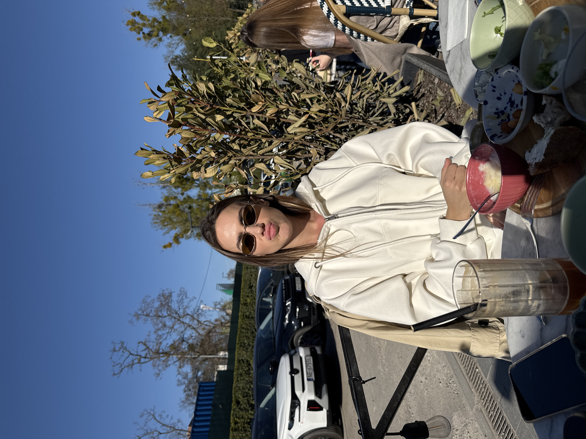 A woman with long brown hair, wearing a white zip-up hoodie and sunglasses, is seated at an outdoor cafe table under a clear blue sky. She looks directly at the camera with her lips slightly pursed, suggesting a posed expression. In front of her on the table is a red bowl containing a light-colored dish (possibly a smoothie or yogurt bowl) with a spoon, and an empty tall glass with a black straw. A dark smartphone rests beside the glass. Several other empty or partially used bowls and plates, along with a piece of bread, are scattered across the marble-patterned tabletop.
Behind the main subject, amidst green foliage, another person with long brown hair is partially visible, also seated at a table. The background reveals parked cars, including a white car and a darker one, suggesting a street or parking area adjacent to the cafe. The white car displays the license plate "NE 6735L". The scene is brightly illuminated by strong, direct sunlight, casting clear shadows and indicating it is daytime, likely an afternoon. Sparse trees and a visible street lamp are also present in the background. The overall setting depicts a casual outdoor dining experience in an urban environment, in what is understood to be Warsaw, Poland.