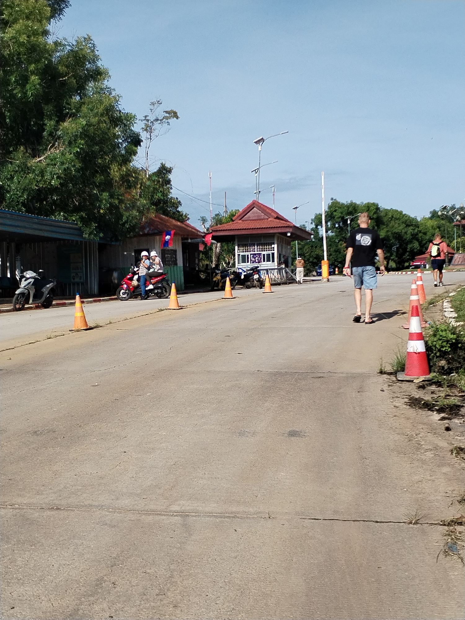 The scene is set on a road in Xayden, Laos. The foreground features a weathered concrete road with cracks and sparse vegetation along the edges. Orange traffic cones line the road. In the midground, several individuals are present. Two people on a red scooter wearing helmets are parked near a small building with a red roof displaying a Laotian flag. Further down the road, a man in a black t-shirt and denim shorts walks away from the camera, while another person with a backpack is walking in the same direction. In the background, there are more buildings, lush green trees, and utility poles. There are also several parked motorcycles. The sky is a clear blue, indicating daytime.