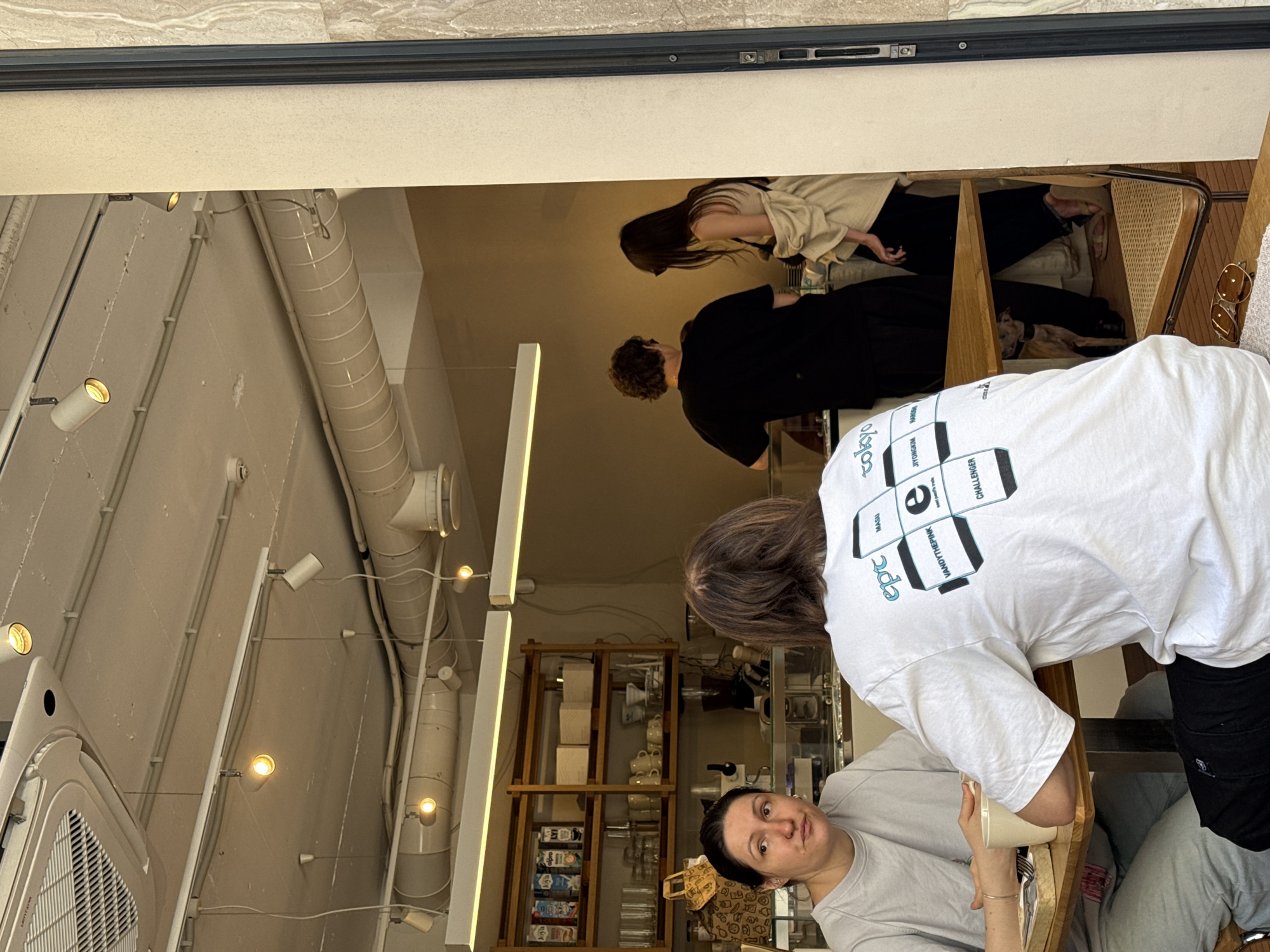 The image captures an indoor scene, likely a cafe, within the Vracar Urban Municipality, Serbia. In the foreground, two individuals are seated at a wooden table. The person on the left, a woman with dark hair pulled back, wears a light grey t-shirt and grey sweatpants. She is looking to her left, with her hands resting on the table next to a mug. The person on the right has their back to the camera, wearing a white t-shirt with graphics on the back and black pants. Their hands are clasped on the table.
In the background, two other individuals are visible. A person wearing a black t-shirt stands behind a counter or service area, and a person with long dark hair, wearing a beige jacket over a light-colored top and black pants, is interacting with them. A small dog is partially visible beneath the table, near the feet of the people in the background.
The environment appears to be a modern establishment with exposed ventilation systems and track lighting on the ceiling. Shelves stocked with products and glassware are visible in the background, suggesting a retail or service area. The overall lighting is bright and natural.