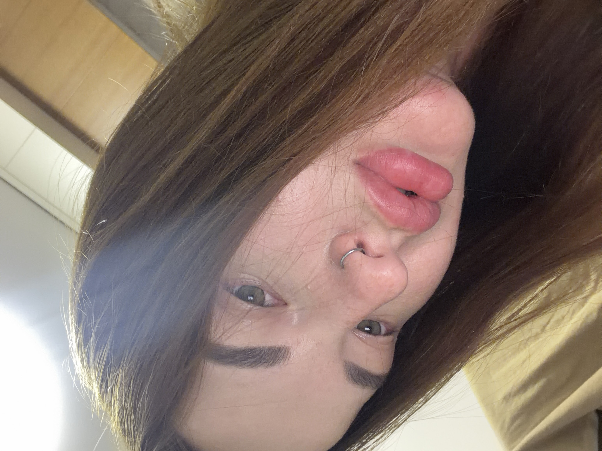 This close-up selfie captures a young person, likely female, with a direct gaze, taken in Krasnodar, Russia. The image is oriented upside down, showing the individual's face from a low angle looking upwards. The subject has medium-brown hair that falls around her face and extends into the background. Her eyes are a striking green or hazel, and her eyebrows are dark and well-defined. Her lips are full and appear a natural pink color. A silver hoop piercing is visible on her right nostril.
The skin tone is light, and there are some subtle highlights on her forehead and nose, possibly from natural oils or lighting. The expression is neutral and direct, without strong overt emotion. The background is soft and out of focus, primarily showing hints of an indoor setting. Elements like a light-colored ceiling with a fixture or vent, a soft brown surface (possibly wood or a wall), and a light-colored fabric (likely clothing) are vaguely visible, largely obscured by the subject's hair. The lighting appears to be artificial and bright, characteristic of an indoor environment, creating some glare on the hair and skin. No visible text is present in the image. The person is posing for the camera, capturing a moment with an intimate and unusual perspective.