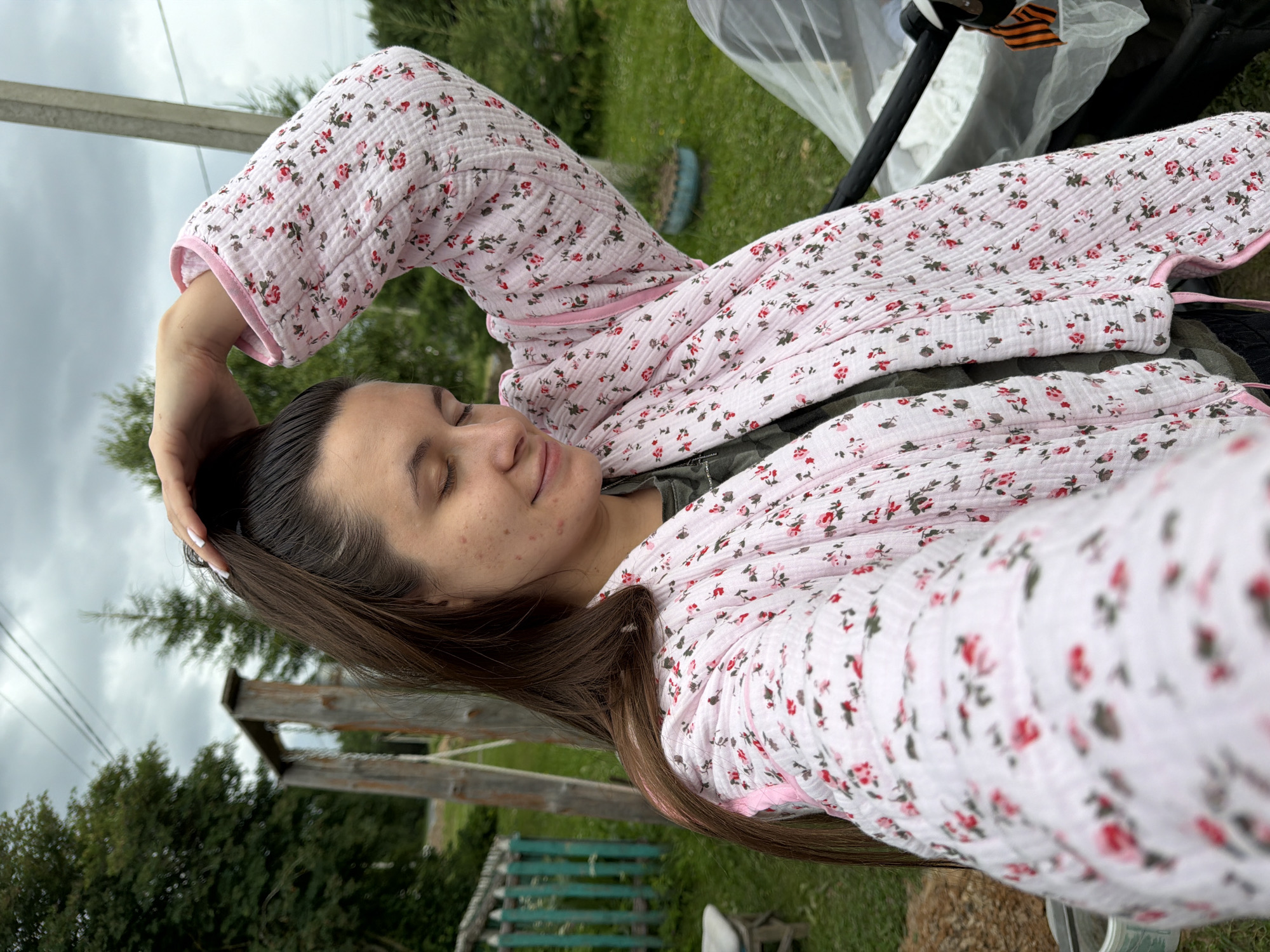 The image shows a young woman taking a selfie outdoors. She has long, dark brown hair and her eyes are closed. She is wearing a light pink, quilted robe with a floral pattern of red and green flowers. Her arm is raised, and she is touching her hair. The background suggests a rural or semi-rural setting with green grass, trees, and a wooden structure that appears to be part of a playground or garden. A stroller or pram is visible in the upper right corner, with what looks like a ribbon with orange and black stripes attached to it. The sky is overcast, indicating a cloudy day. The overall mood seems relaxed and peaceful. The location is identified as Krasny Mayak, Russia. No visible text is present in the image.