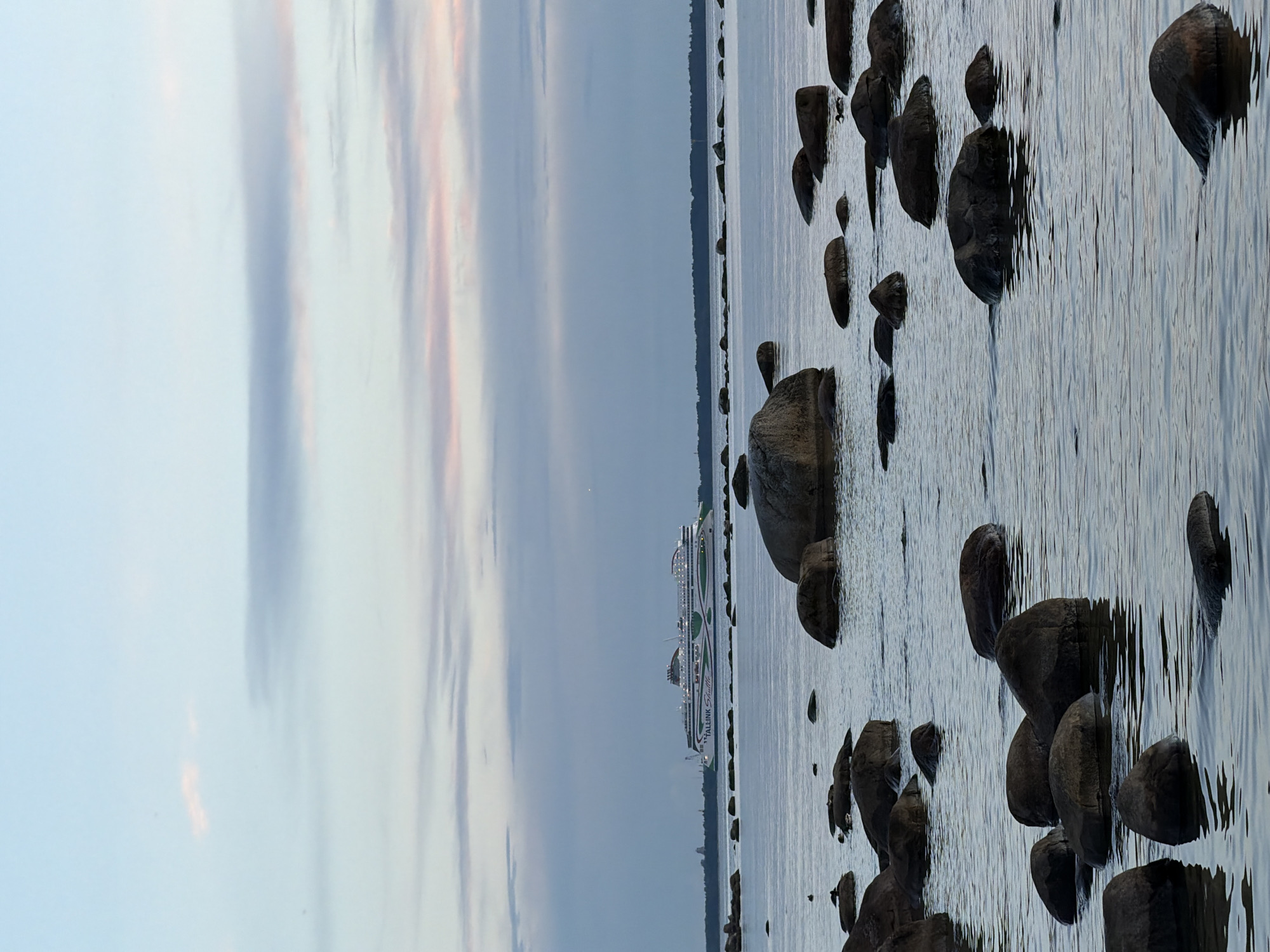 The image depicts a coastal scene with a large passenger ferry in the background, sailing on a calm body of water. The ferry is white with green accents and the text "TALLINK Shuttle" is visible on its side. In the foreground, numerous dark, smooth rocks of varying sizes are partially submerged in the water, creating a natural, rugged shoreline. The sky is overcast with soft pastel hues of blue, pink, and grey, suggesting either dawn or dusk. The overall environment is serene and natural, characteristic of a coastal area. The scene is located in Haabneeme alevik, Estonia, as indicated.