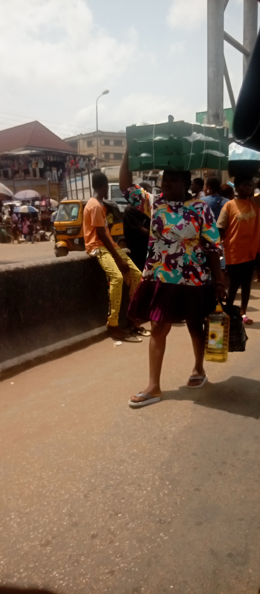 The image shows an outdoor scene in Onitsha, Nigeria. A woman wearing a colorful short-sleeved top and a short purple skirt walks toward the camera, carrying a stack of wrapped green rectangular objects on her head and a large plastic bottle of cooking oil in her right hand. She is wearing flip-flops. To her left, a young man in a yellow patterned outfit sits on a low concrete wall. In the background, there is a bustling marketplace with many people, stalls, and yellow tricycle taxis. There are buildings, and a cloudy sky can be seen.