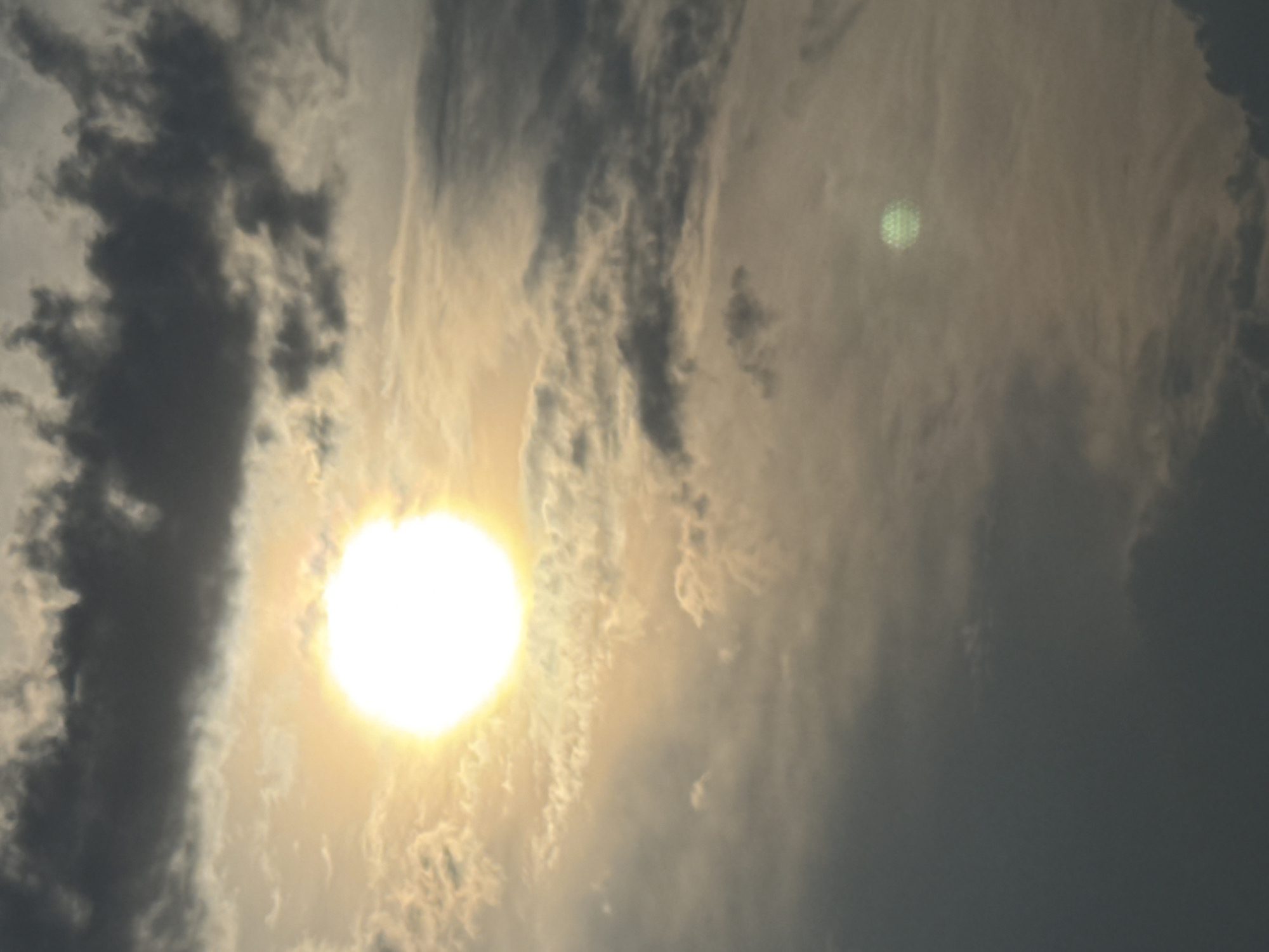 The image depicts a sky filled with clouds and the sun. The sun, a bright white orb, is positioned in the upper left quadrant of the image, partially obscured by thin, wispy clouds. In the lower right quadrant, a smaller, faintly green, disc-shaped object is visible, also surrounded by clouds. The clouds vary in density and color, ranging from dark gray to lighter, hazy tones. The overall impression is of a cloudy day with the sun shining through. The location is identified as Moscow, Russia.