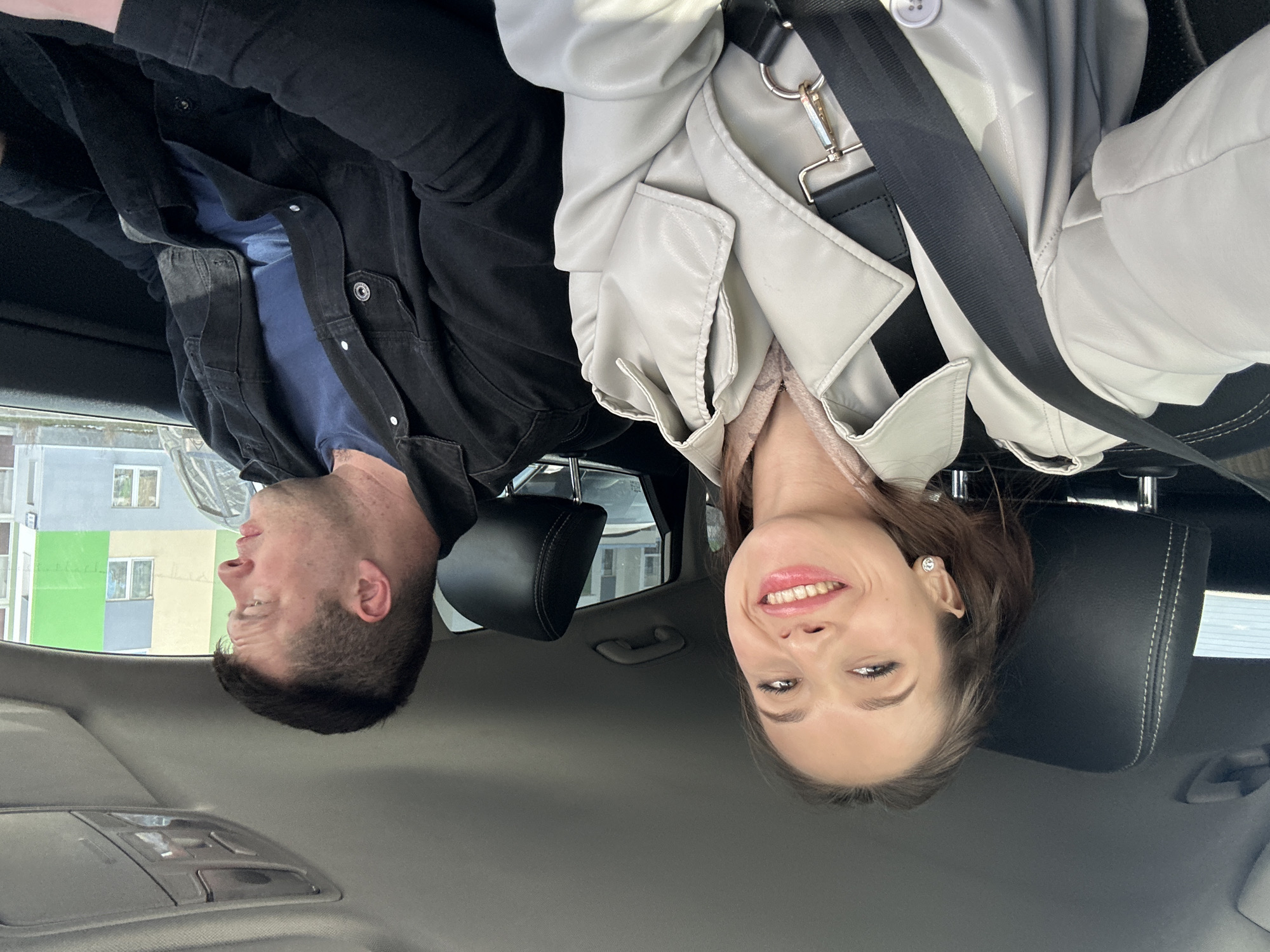 Inside a car, a man and a woman are visible, both wearing seatbelts. The man is in a black jacket over a blue shirt. The woman is in a light gray jacket. Outside the car window, a multi-colored building is visible. Given the context, this image was likely taken in Yekaterinburg, Russia.