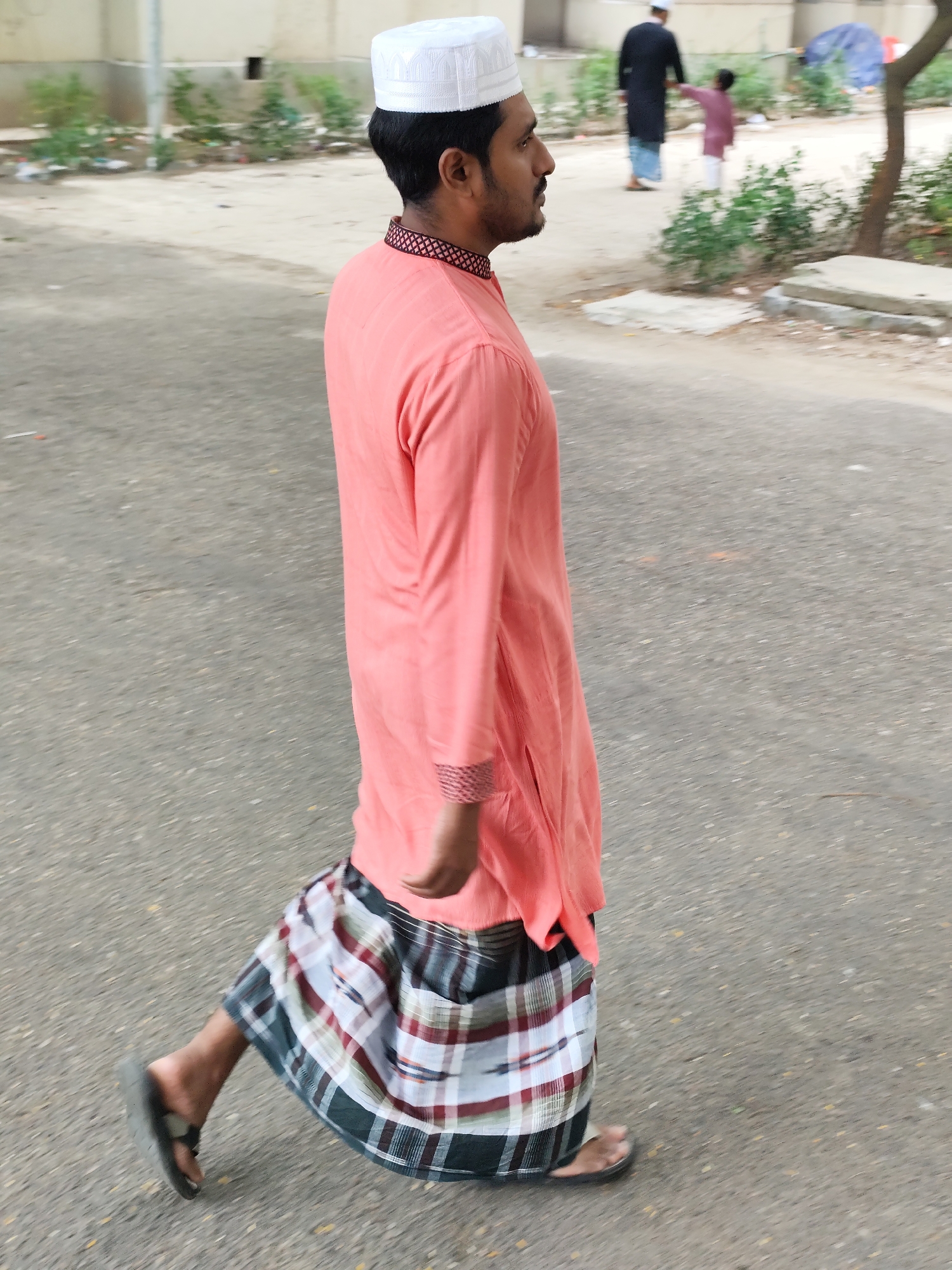 A man wearing a pink kurta, a white skullcap, and a checkered lungi walks on an asphalt road. He is wearing sandals. In the background, a man in a dark garment and a child in a pink shirt are walking away from the camera. The environment is urban, with a few trees and buildings visible. There is some debris on the side of the road. The lighting suggests it is daytime. The image appears to be taken in Dhaka, Bangladesh.