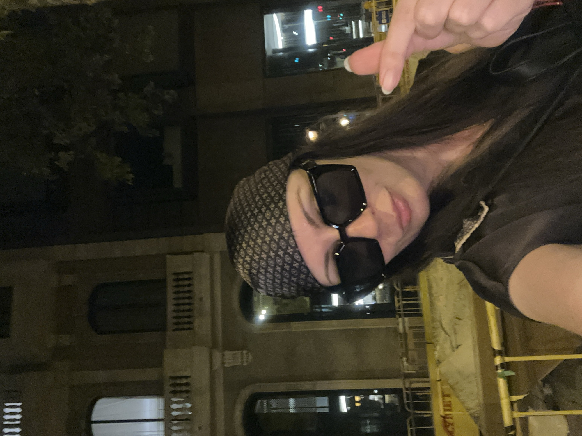 The image captures a selfie of a woman at night in Barcelona, Spain. She is wearing a bandana, large square sunglasses, and a black top. Her right hand is raised, making a finger heart gesture. Behind her, a dimly lit building with arched windows and ornate balconies is visible, consistent with the architecture found in Barcelona's historic districts. The background also shows streetlights and construction barriers, indicating an urban setting.