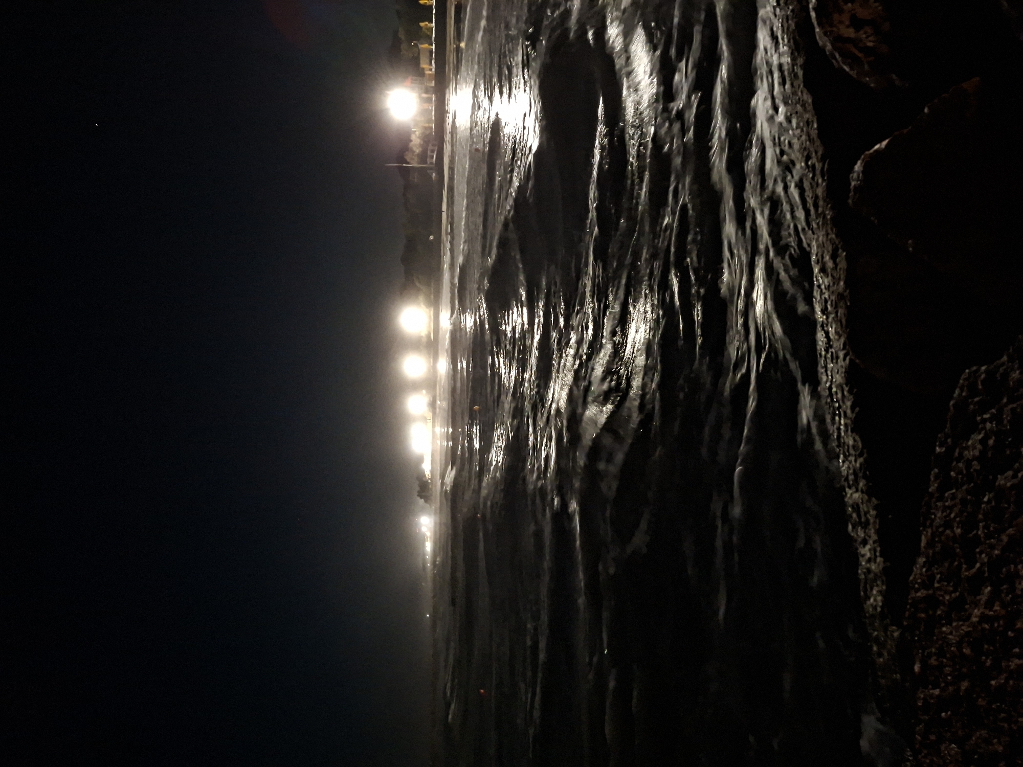 A night view of the Adriatic Sea from the coast of Caorle, Italy. The foreground shows the dark, choppy surface of the sea, with some rocks visible on the right side of the frame. In the background, there are several bright lights illuminating the coastline and some trees. The sky is dark and clear, with a single star visible. The image captures a serene and tranquil atmosphere, with the gentle sounds of the waves and the distant lights creating a sense of peace and calm.